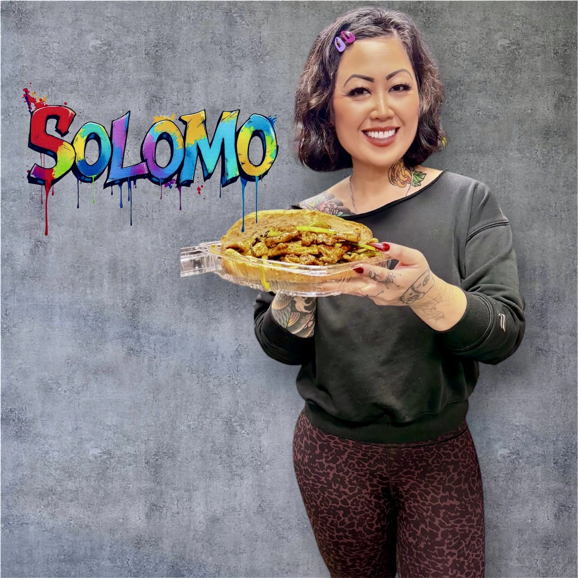 A woman with short curly hair, tattoos, and a big smile holding a dish of food, standing against a textured gray wall with colorful graffiti that reads 'SOLOMO'.