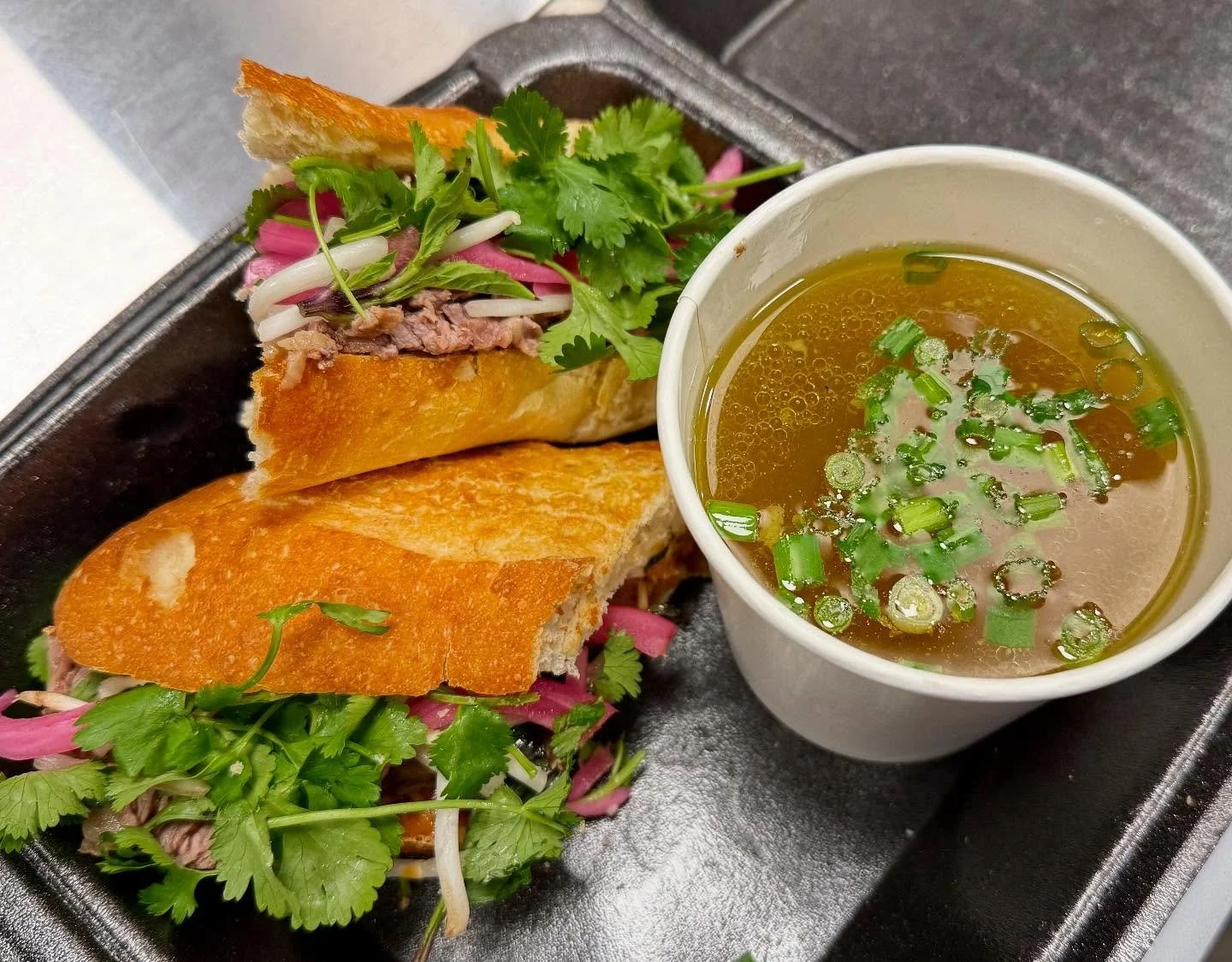Vietnamese banh mi sandwich with sliced bread, grilled meat, fresh herbs, pickled vegetables, and a side of hot broth garnished with chopped green onions