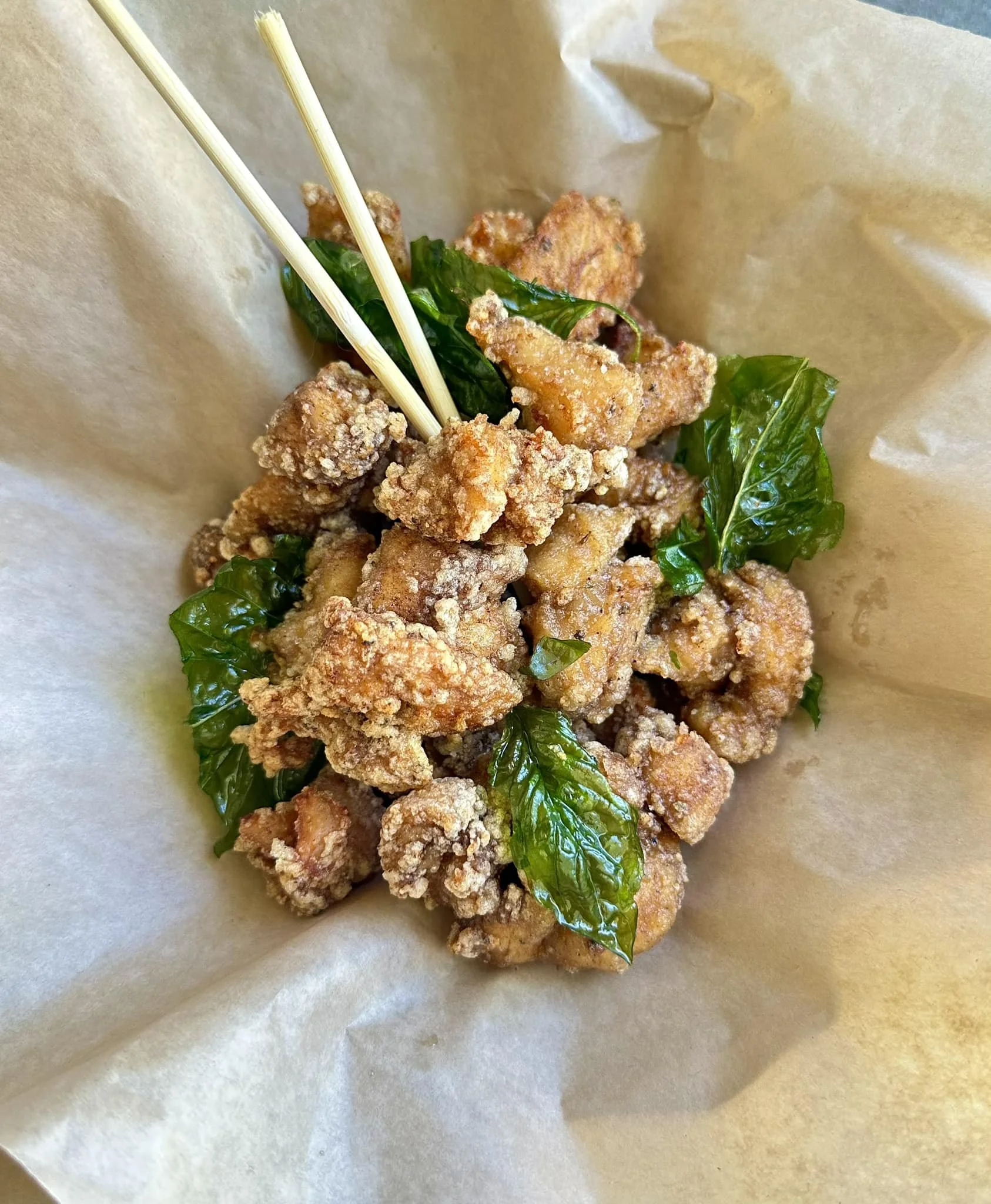 Close-up of fried chicken pieces with green herb leaves and two skewers on a paper-lined surface.