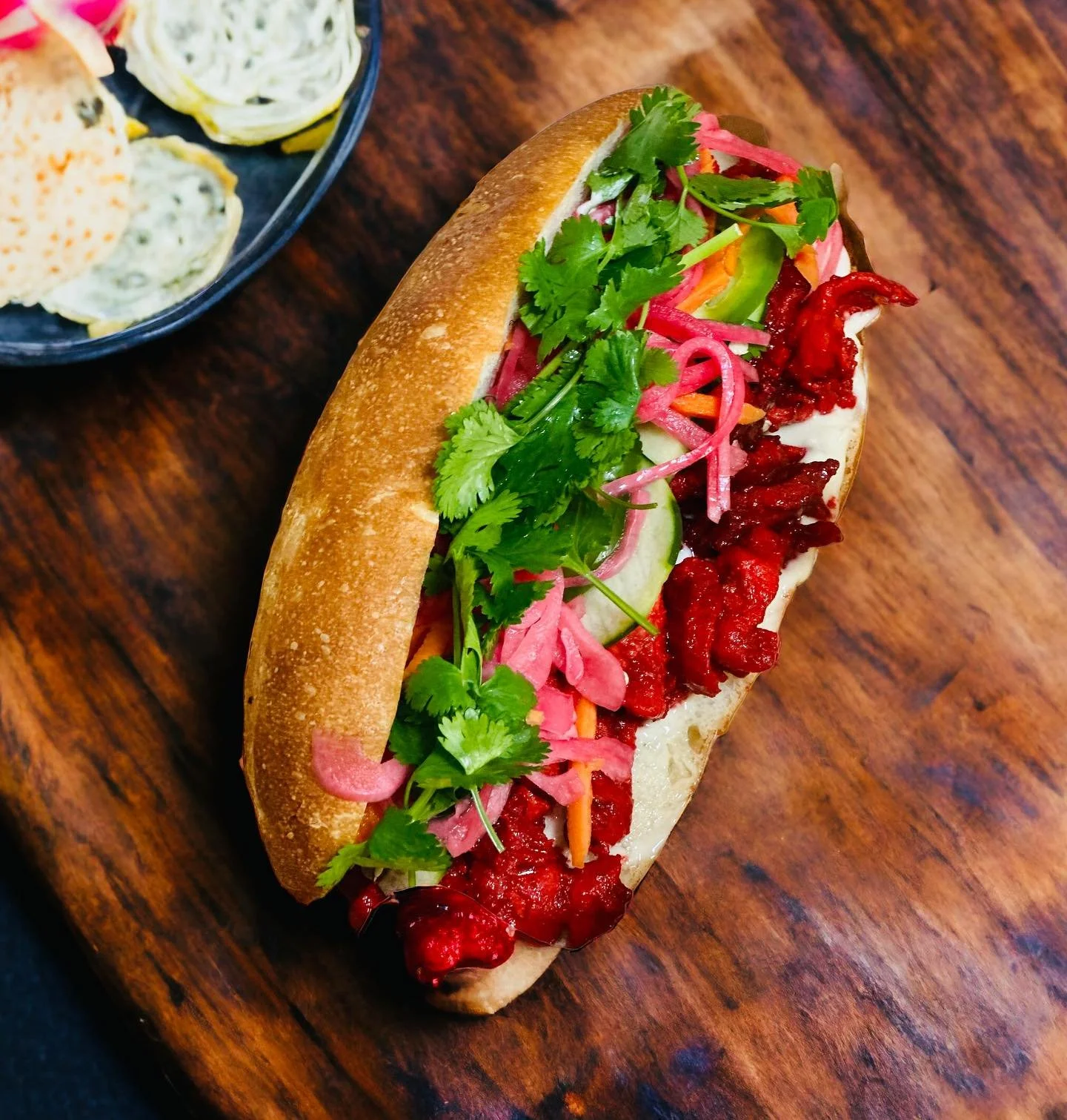 A hotdog topped with cilantro, pickled vegetables, and spicy sauce on a toasted bun, placed on a wooden table.
