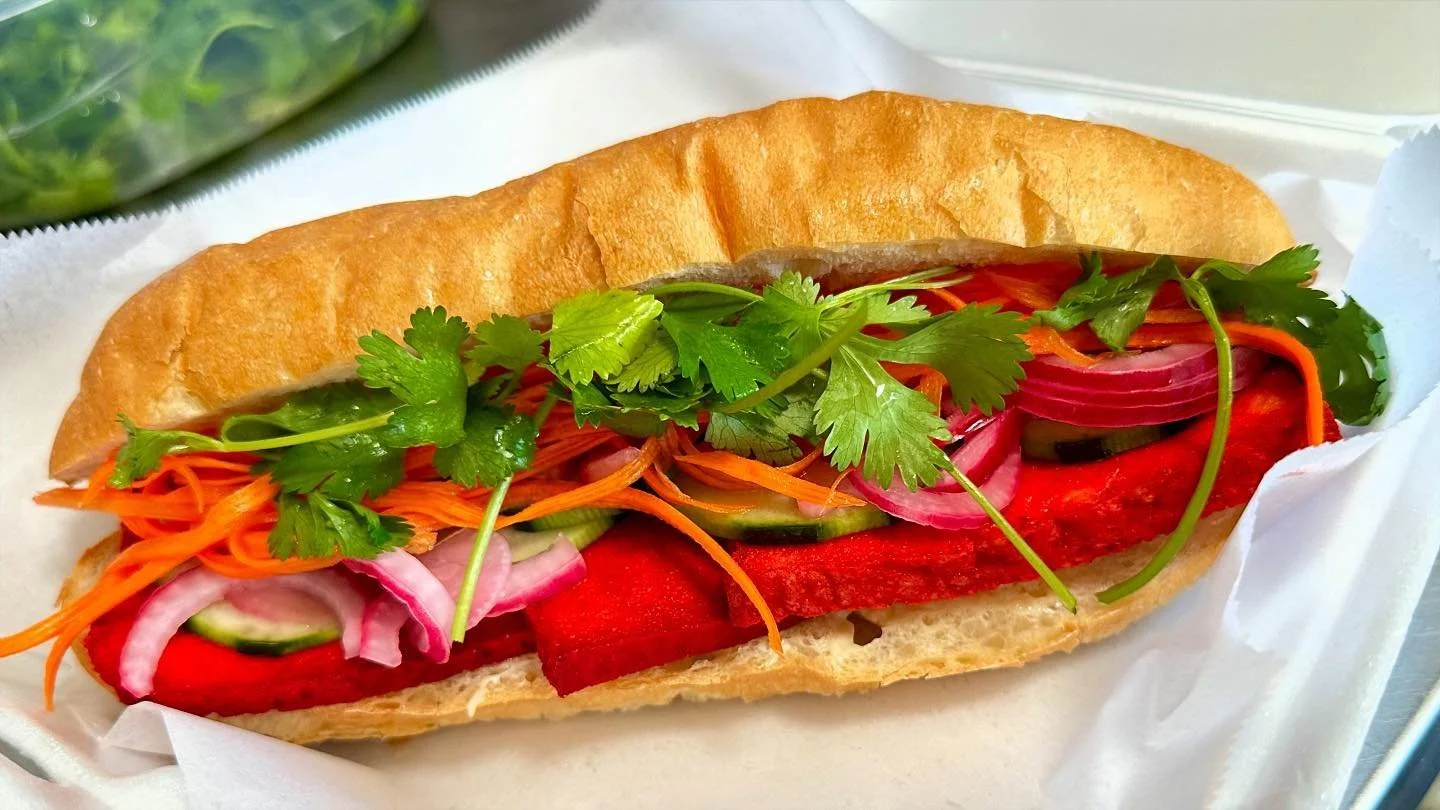 Close-up of a sandwich filled with carrots, cucumbers, red onions, cilantro, and red peppers in a hot dog bun on white paper