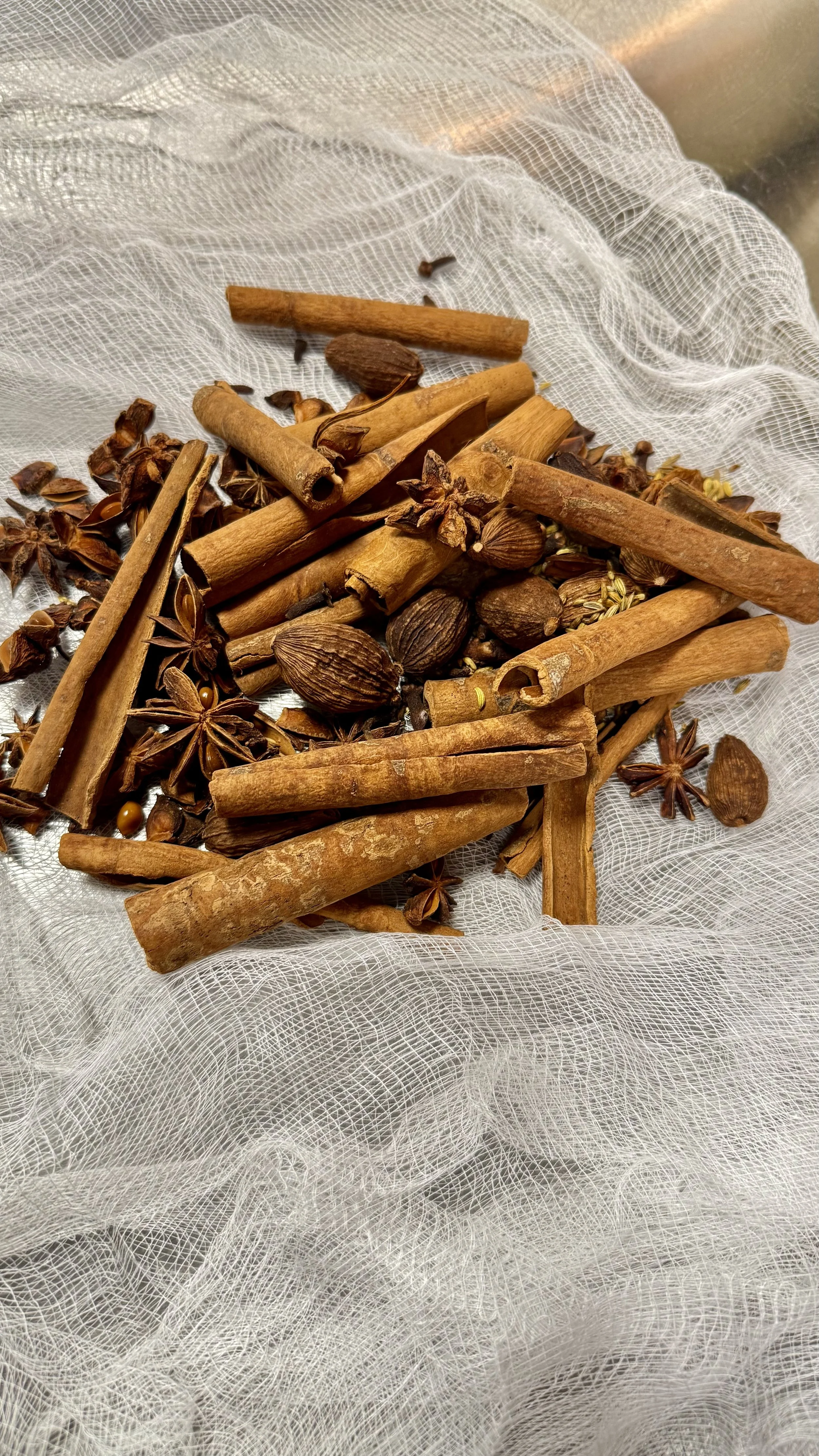 A collection of cinnamon sticks, star anise, allspice berries, and other whole spices on a white mesh fabric.