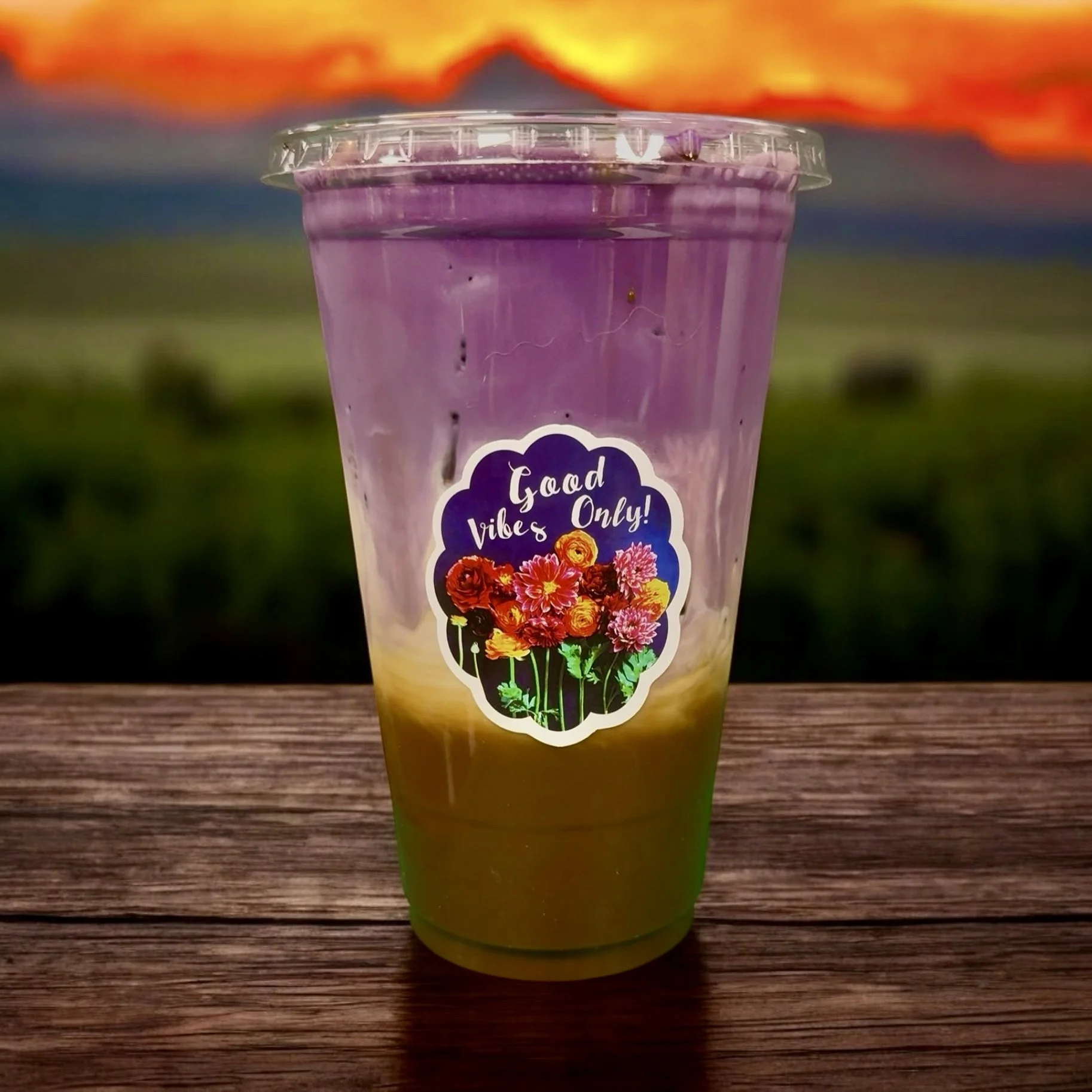 A clear plastic cup with a lid, containing a layered beverage with purple on top, yellow in the middle, and green at the bottom. The cup has a sticker that reads 'Good Vibes Only!' with a colorful bouquet of flowers. Ube Vietnamese coffee