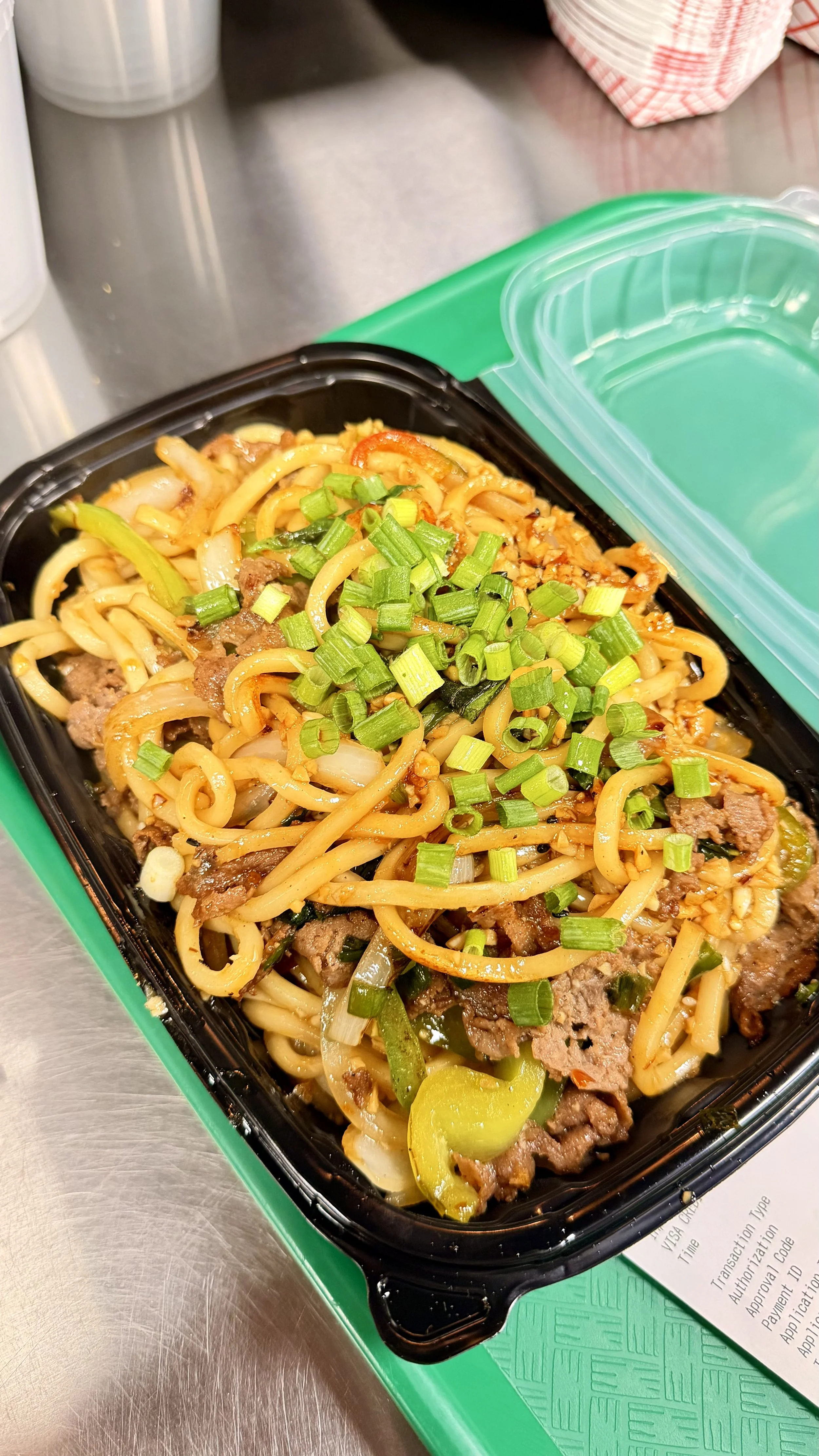 Takeout container filled with stir-fried noodles, beef, green onions, and vegetables on a green tray.