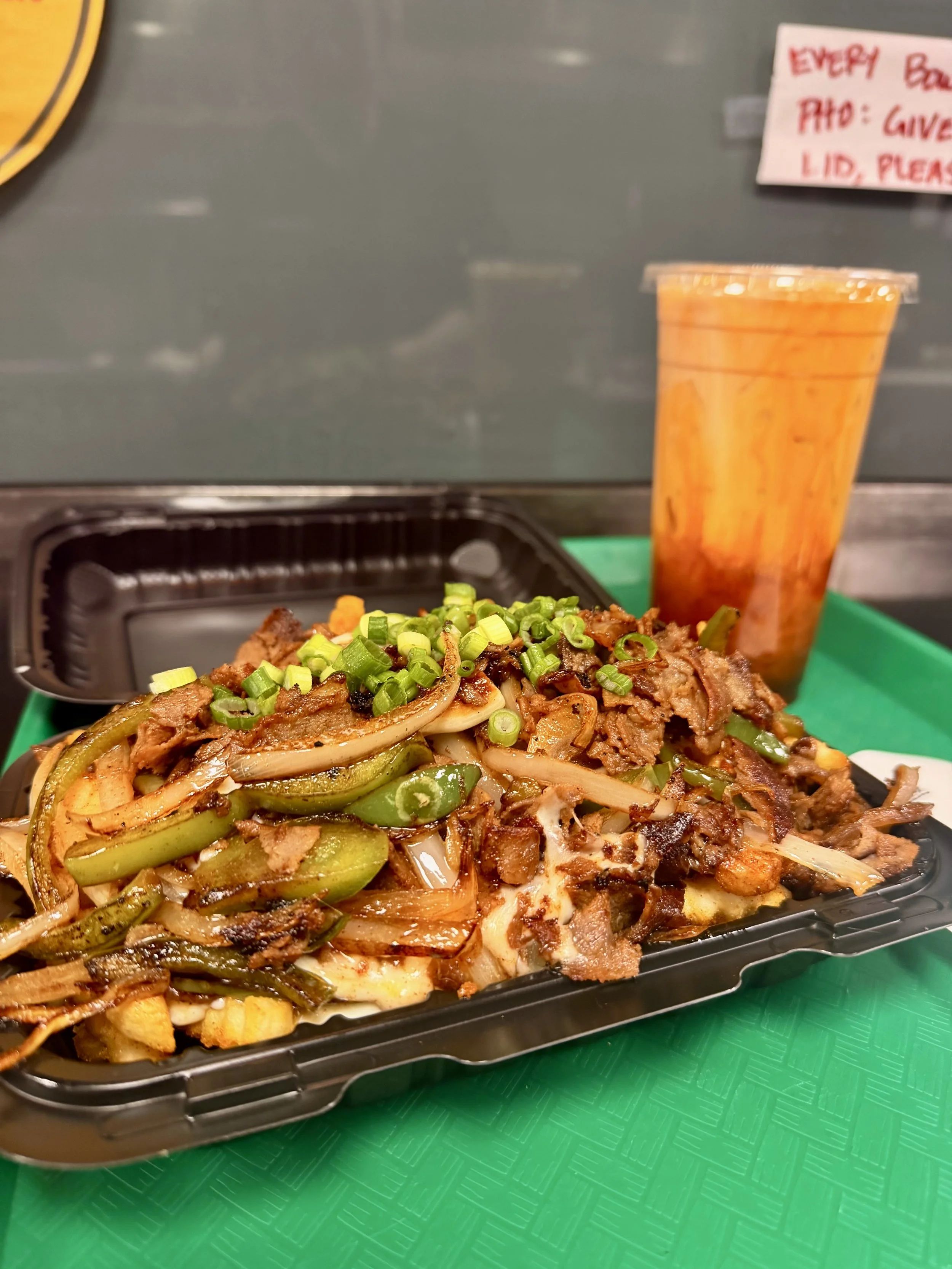A meal on a black plastic tray with chopped green onions on top. A large drink in a clear plastic cup is to the right of the tray. The background is a green surface and a blurry sign with red handwriting.
