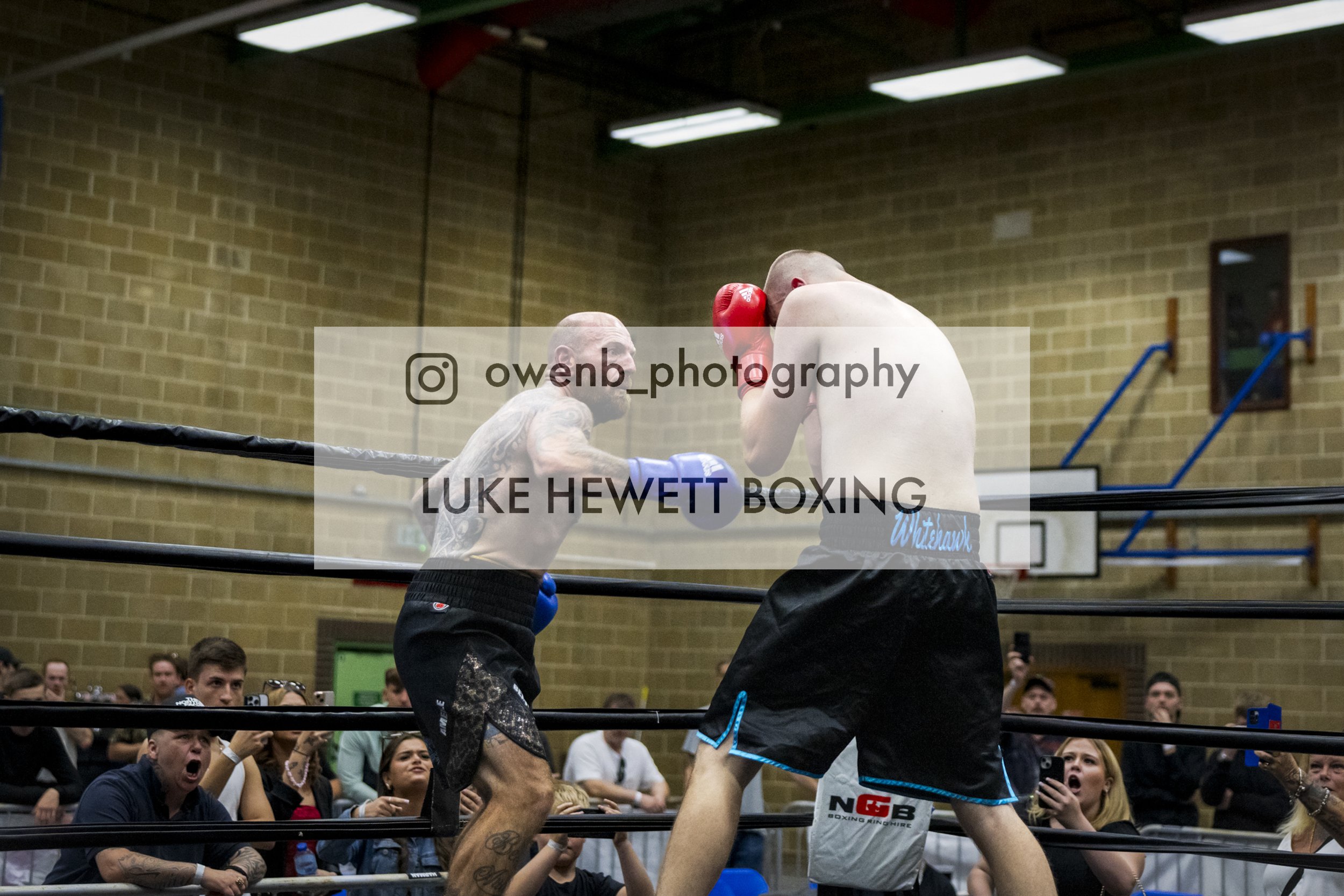 Two men engaged in a boxing match inside a boxing ring, surrounded by an audience, with a brick wall in the background.