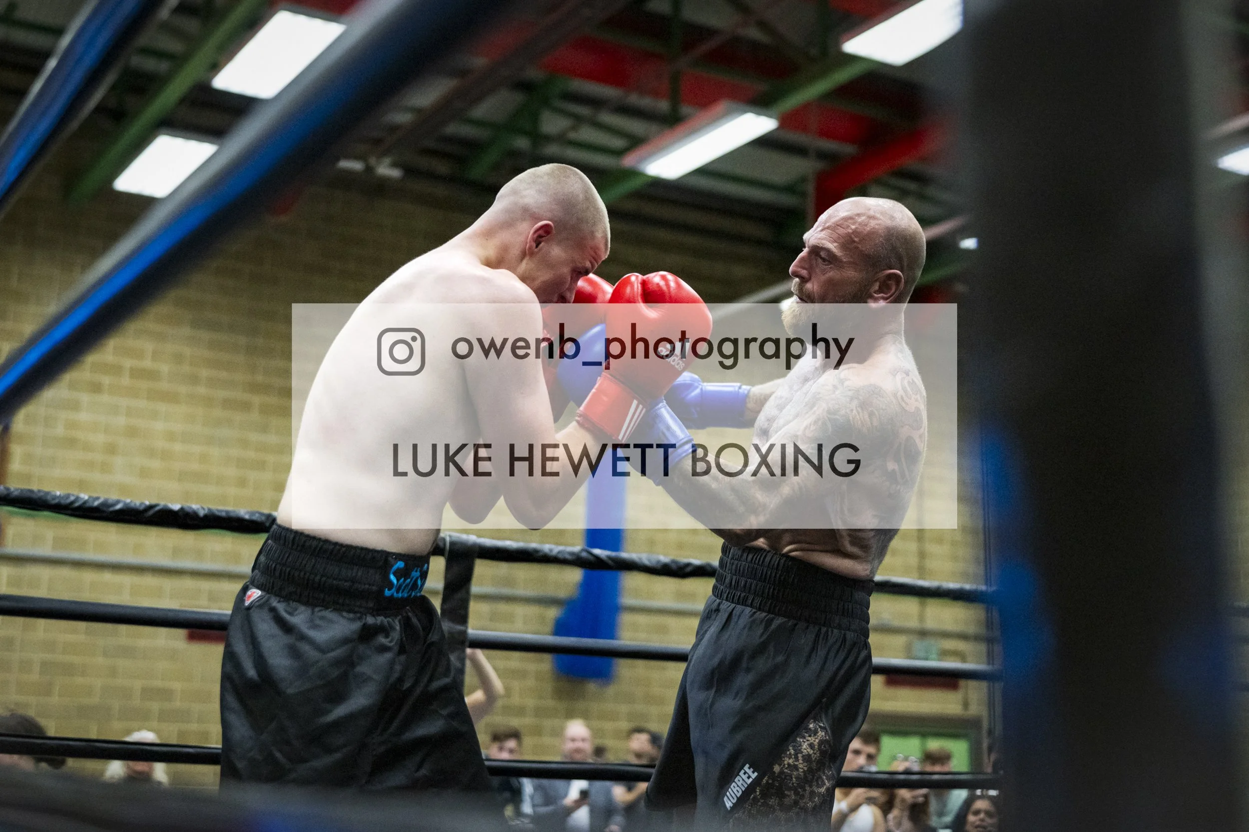 Two shirtless male boxers wearing red and blue gloves in a boxing ring, practicing a clinch while facing each other, with an audience watching in the background.