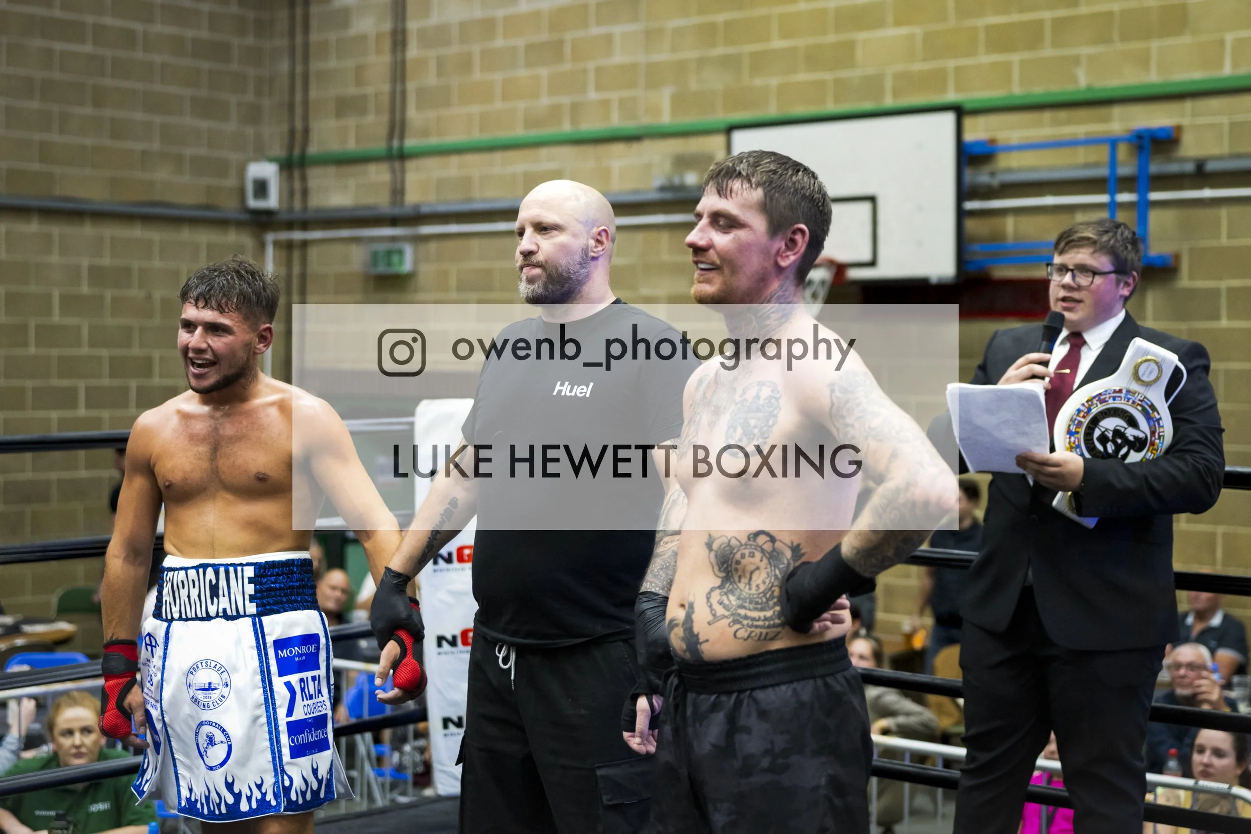 Boxers in a ring after a match, one shirtless with blue and white shorts and the other in black shorts with tattoos, with an announcer in a suit holding a microphone and a championship belt.