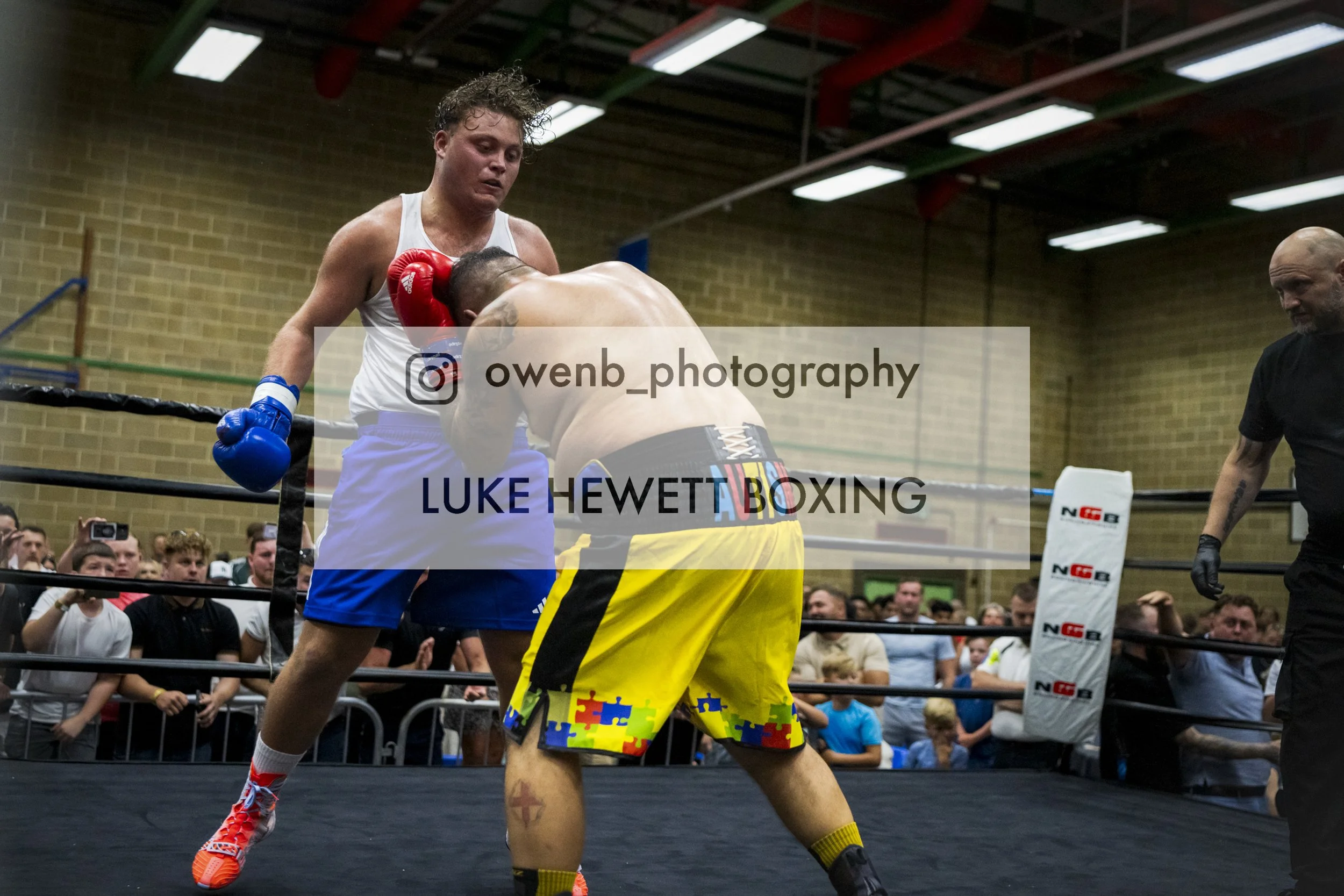 Two boxers competing in a match inside a boxing ring, with a crowd watching in the background, one in white shorts and red gloves, the other in yellow shorts with a multicolor pattern and black gloves, with a referee on the side.