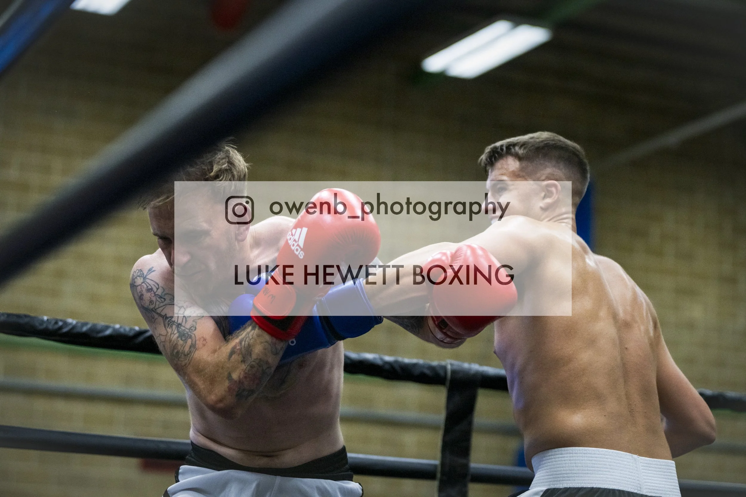 Two shirtless men with boxing gloves engaged in a boxing match inside a gym, with a boxing ring and wooden ceiling in the background.