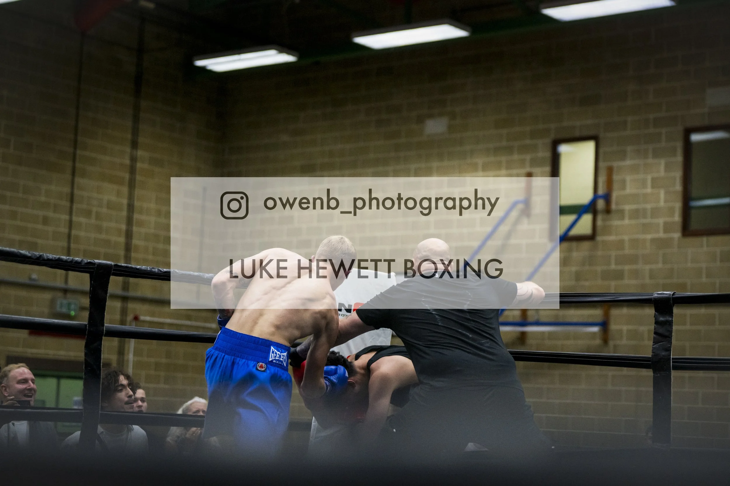 Boxing match in a gymnasium with two fighters in the ring, one wearing blue shorts and the other in black, engaged in a close exchange of punches. Several spectators are watching and cheering. The scene is illuminated by overhead fluorescent lights, 