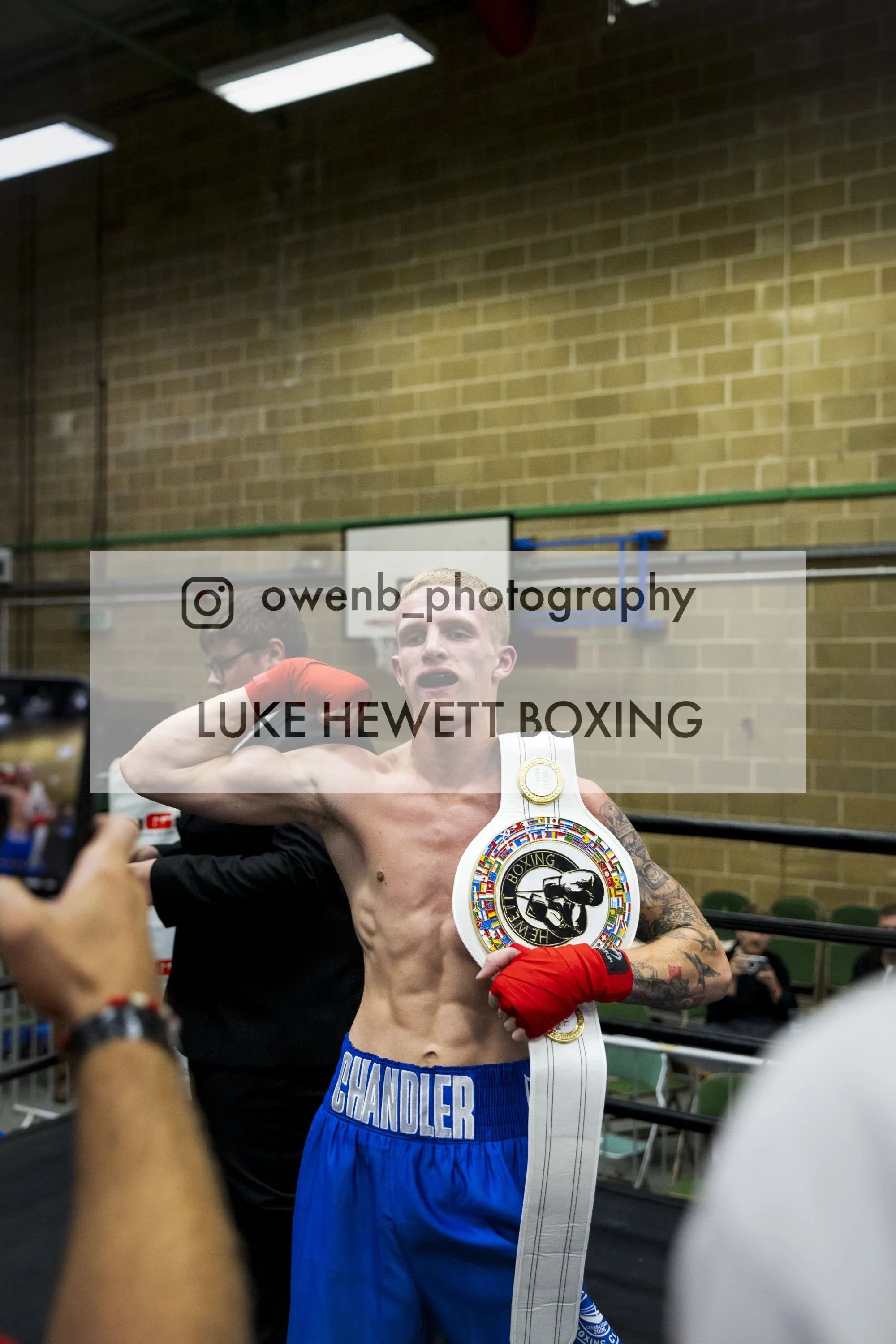 A shirtless boxer with a championship belt over his shoulder, showing his toned physique and tattoos, in a gym with brick walls and green chairs.