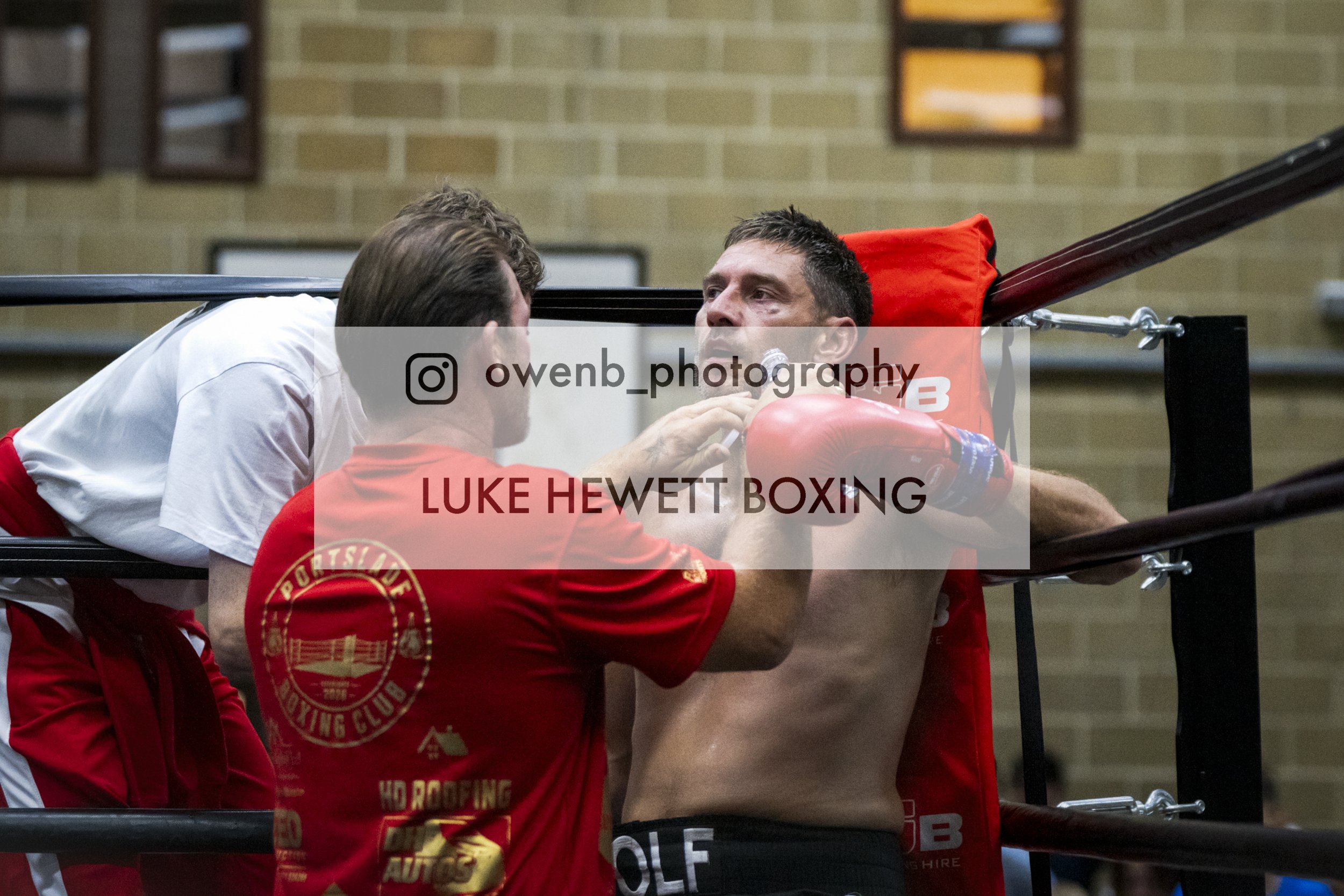Boxer Luke Hewitt receiving advice from a coach during training in a boxing ring, with another person in red training attire nearby.