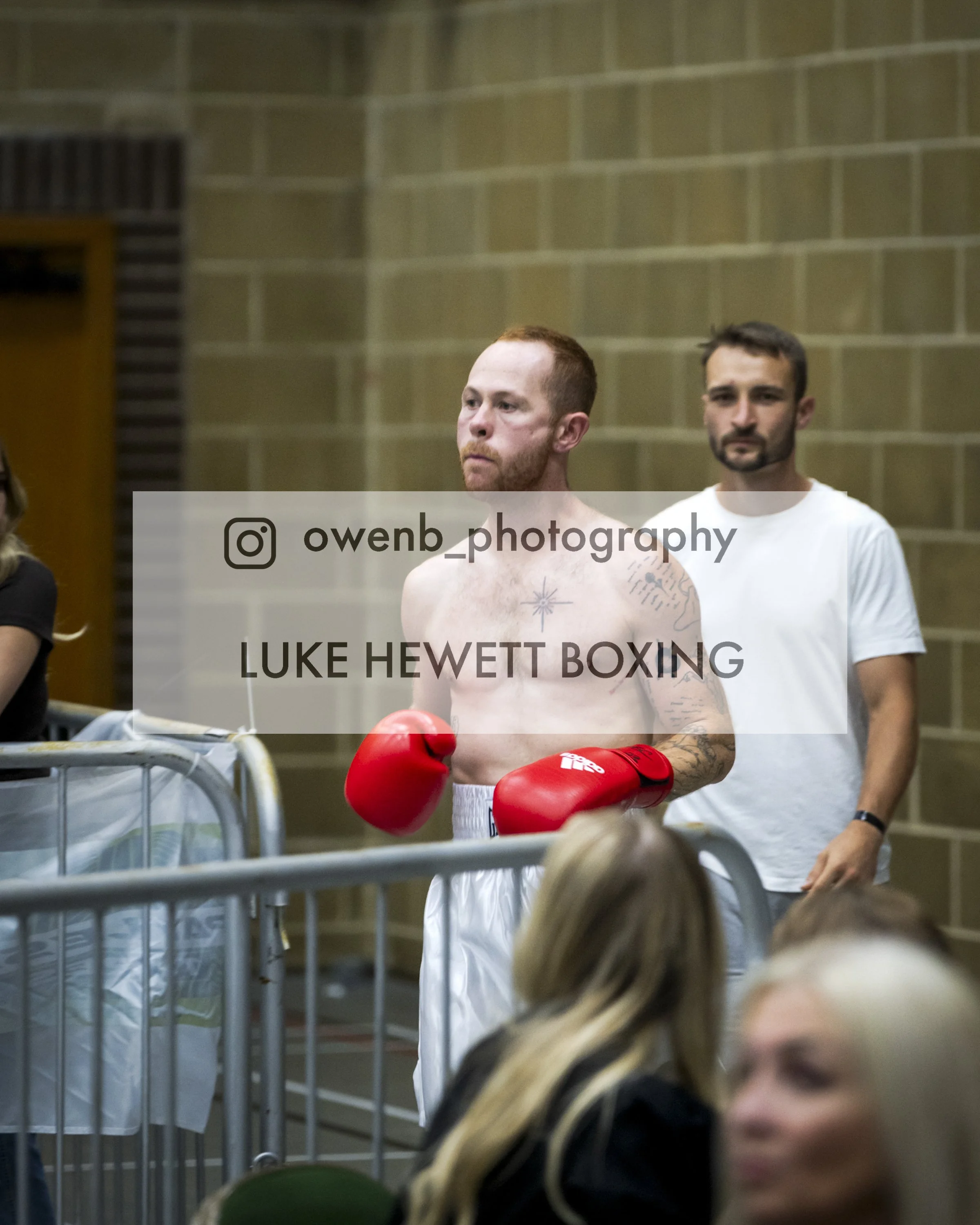 A shirtless man with red hair and tattoos, wearing red boxing gloves, stands in a boxing gymnasium with a beige brick wall behind him. Another man with a beard and wearing a white t-shirt stands nearby. There are people and equipment in the foregroun