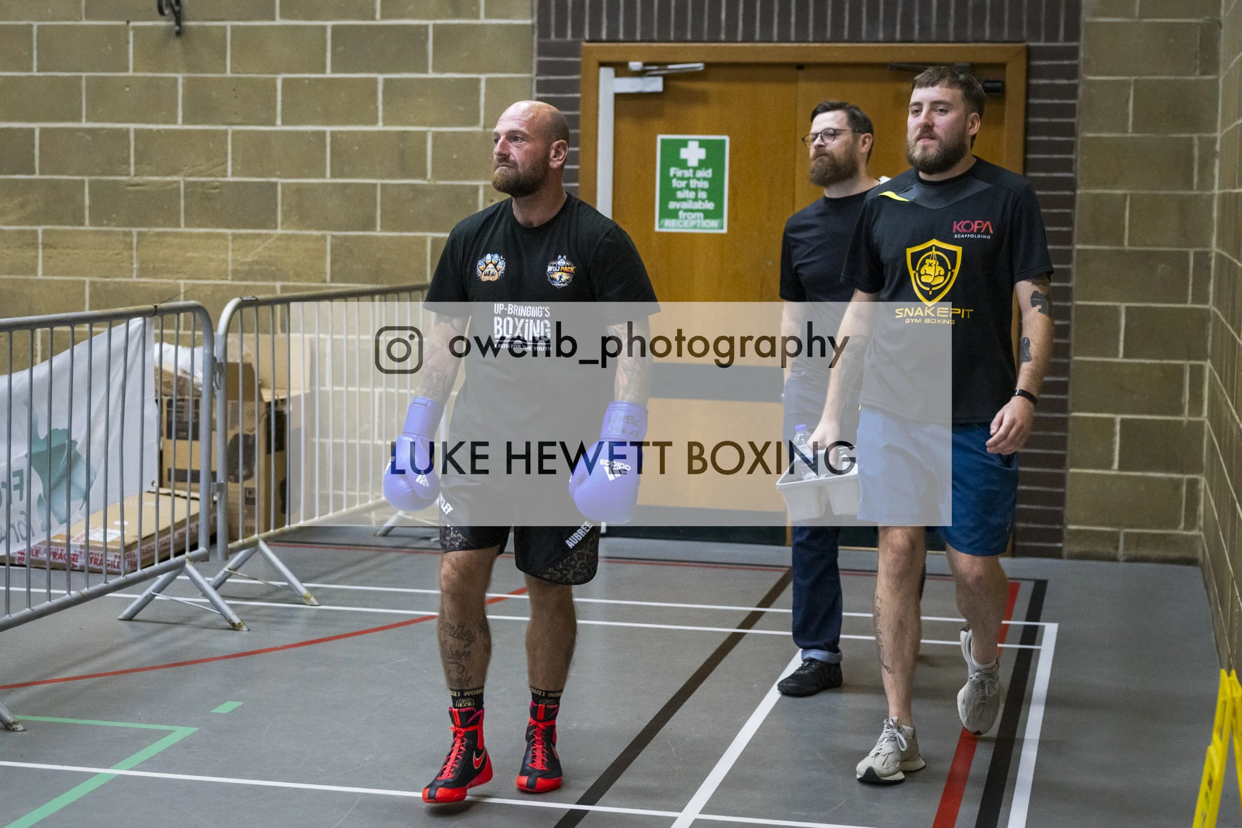 A boxer with a shaved head and tattoos walks in boxing gloves, followed by two men, one in glasses, in casual clothing, walking in an indoor gymnasium. Bell markers and a first aid sign are visible in the background.