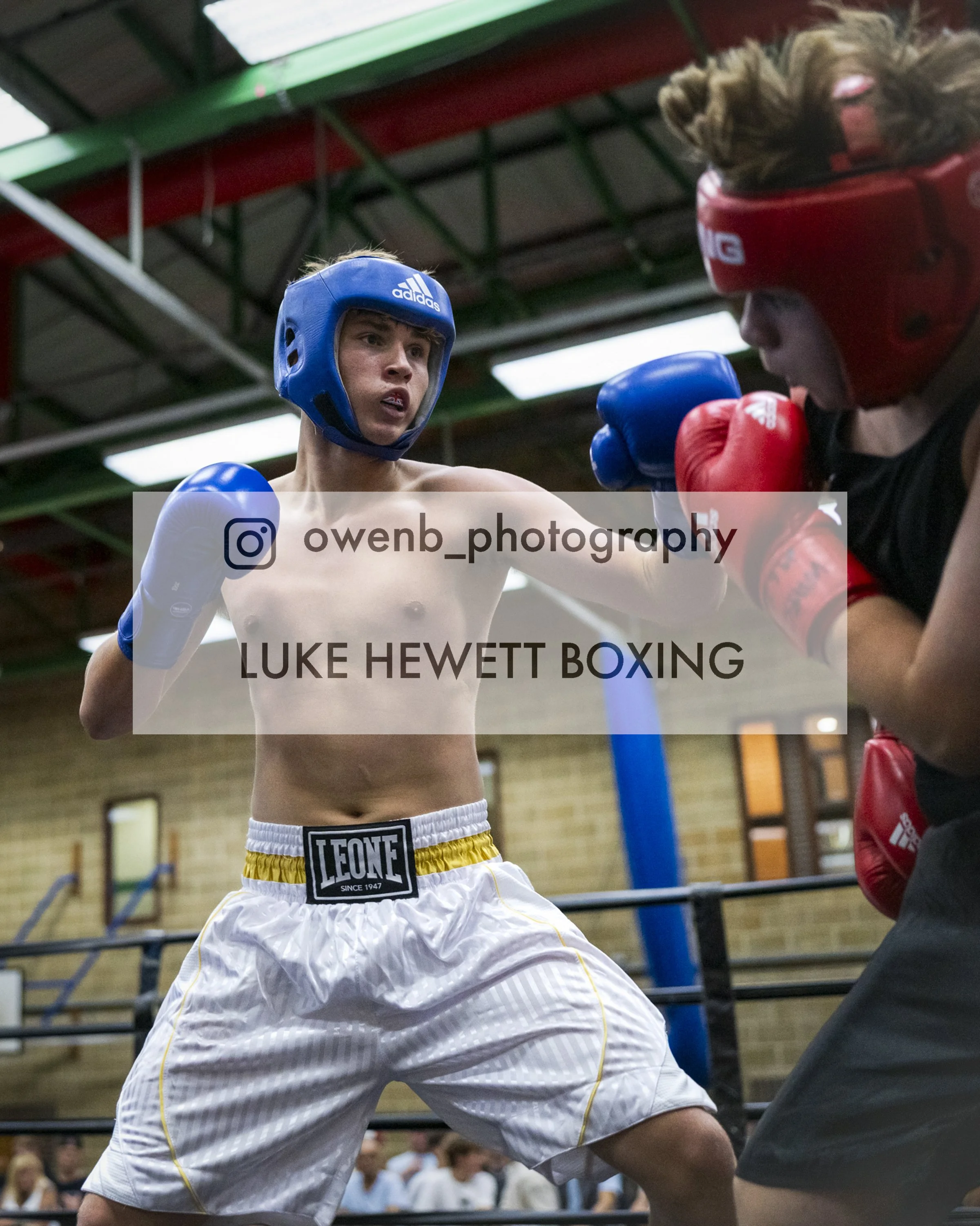 Two young male boxers wearing protective headgear and gloves sparring in a boxing gym. One is dressed in yellow and white shorts, the other in black and red gear. They are engaged in a close fight with focused expressions.