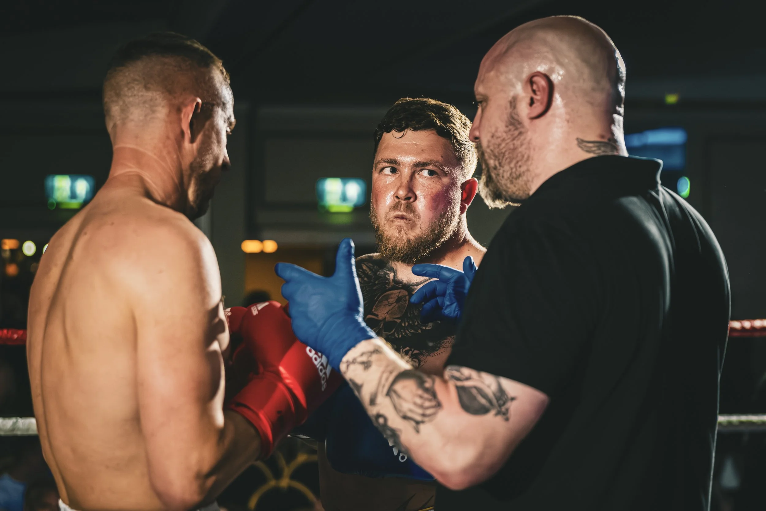 Two shirtless men, one with tattoos and one with a beard, face off in a boxing ring while a third man, with a shaved head and a tattoo on his neck, talks to them, wearing blue gloves and a black shirt. The setting appears intense and serious.