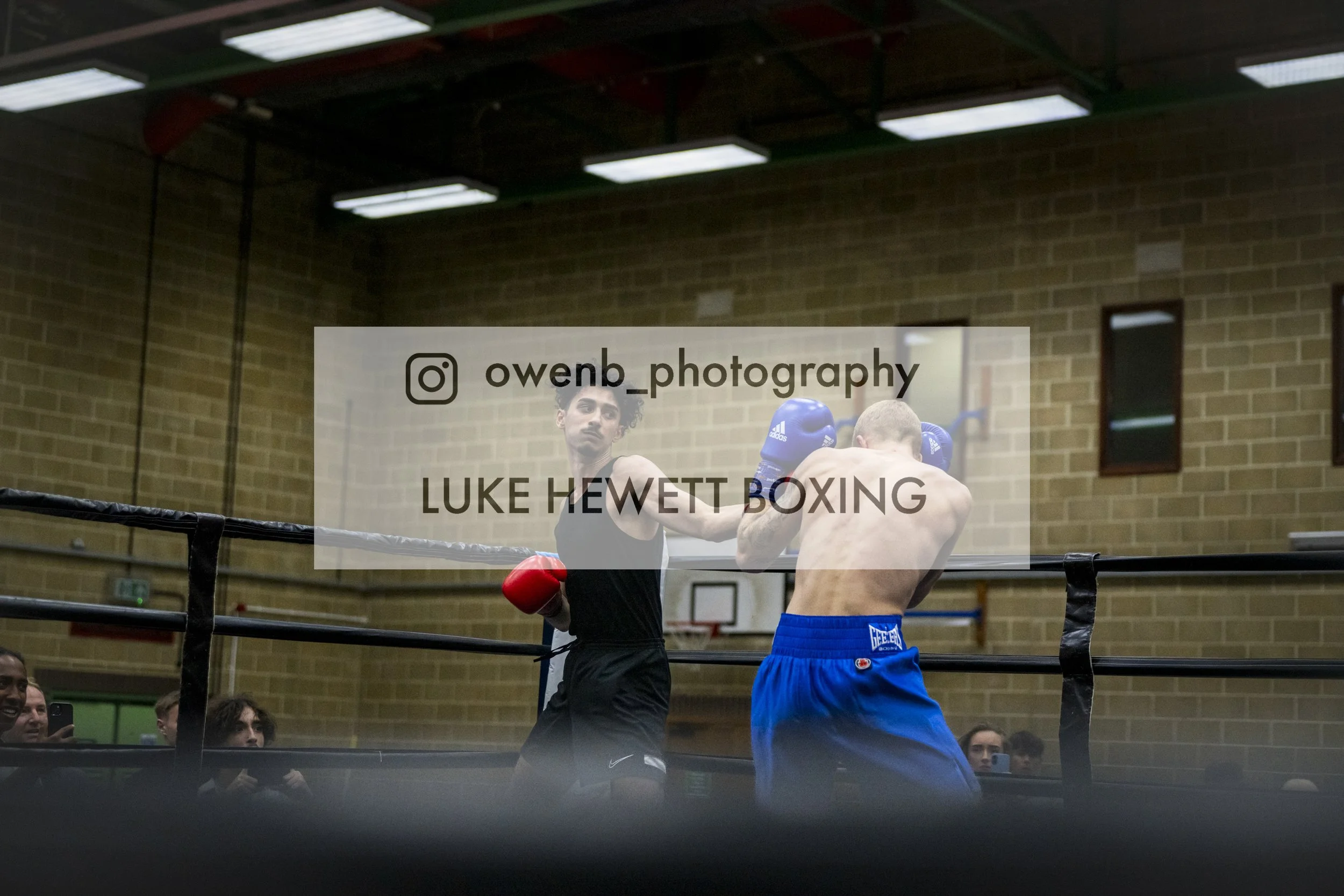 Two boxers are engaged in a boxing match inside a gymnasium, with one boxer wearing red gloves and black shorts, and the other wearing blue gloves and blue shorts. They are fighting near the ropes of the boxing ring, with spectators watching in the b