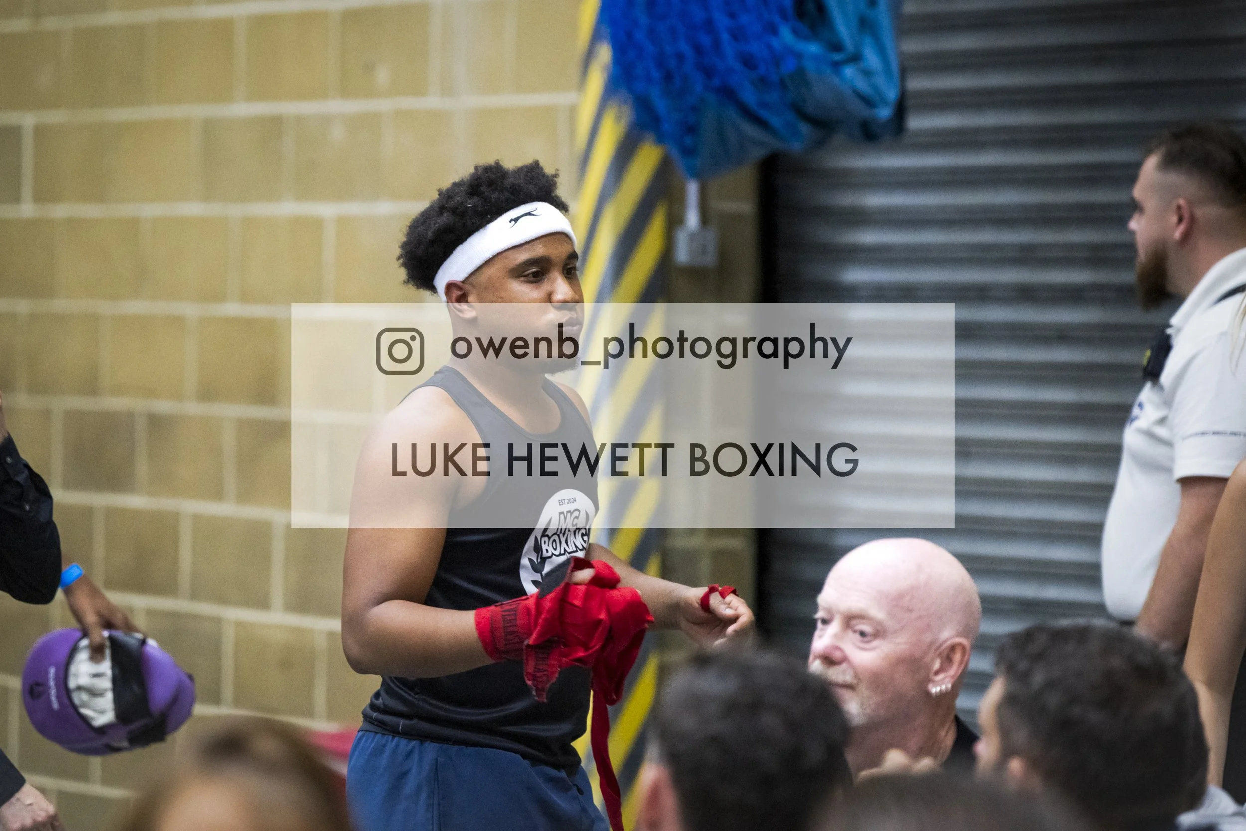 A young female boxer with a white headband and red gloves stands in a gym, holding her gloves, as she interacts with individuals including a bald man with a beard and an earring, with gym equipment and a group of people visible in the background.