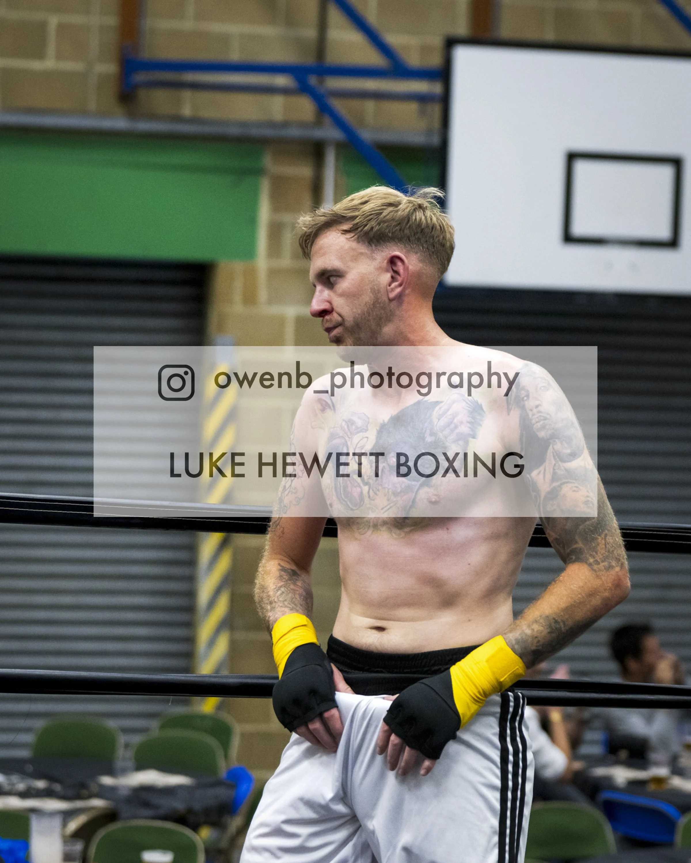 A shirtless man with tattoos on his chest and arms, wearing yellow and black boxing gloves, standing in a gym with boxing rings in the background.