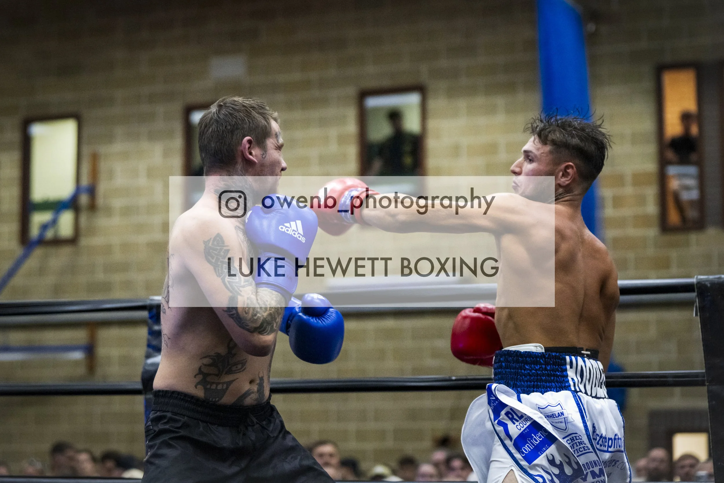 Two shirtless male boxers with gloves engaged in a boxing match inside a gym. One has tattoos and is throwing a punch at the other, who is wearing blue and white shorts. The background shows a brick wall and mirrors.