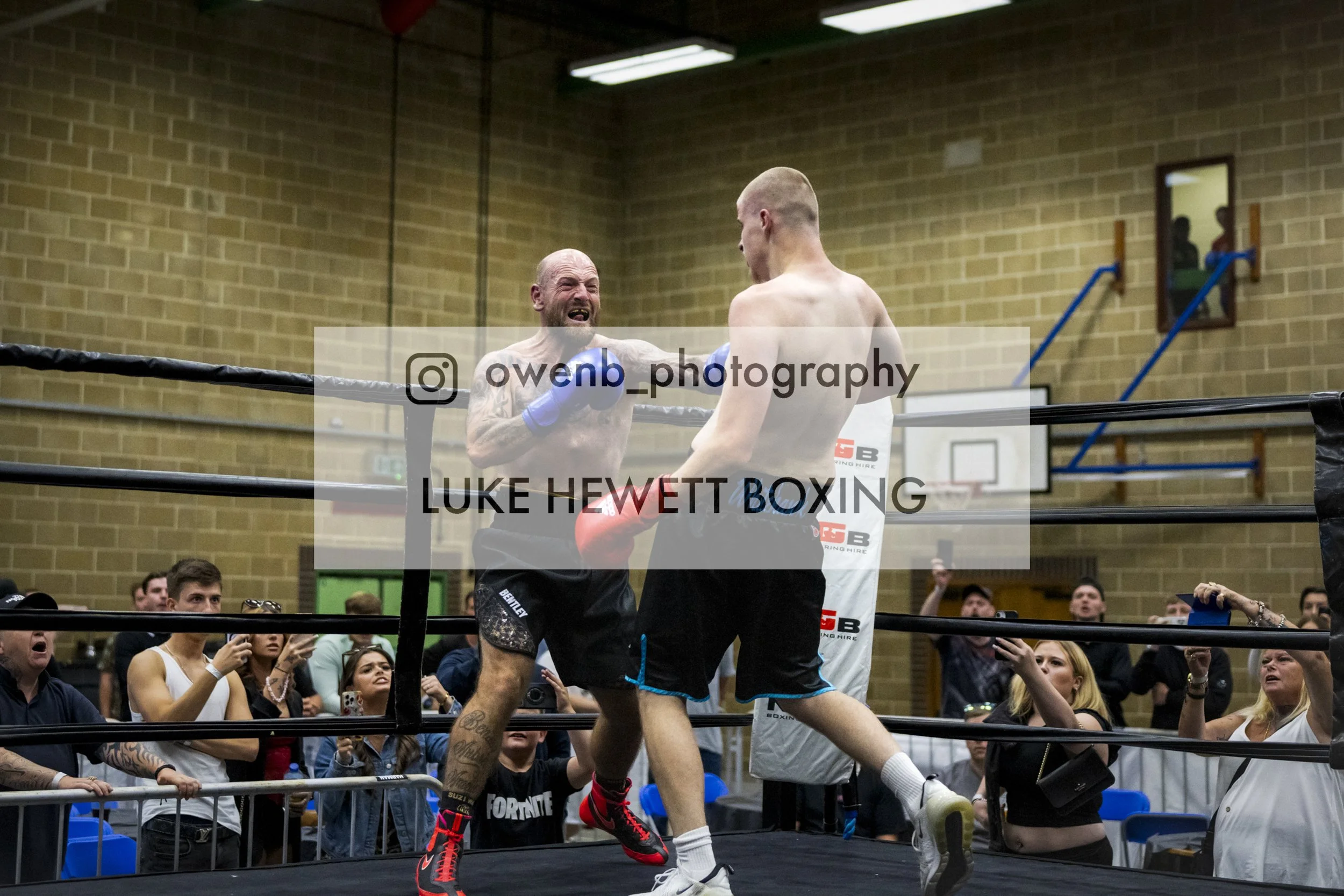 Two male boxers in a boxing ring, one with a beard and tattoos, the other with a shaved head, are fighting with the crowd watching around them. The boxer with tattoos is grimacing while the other is facing away. The audience appears excited and engag