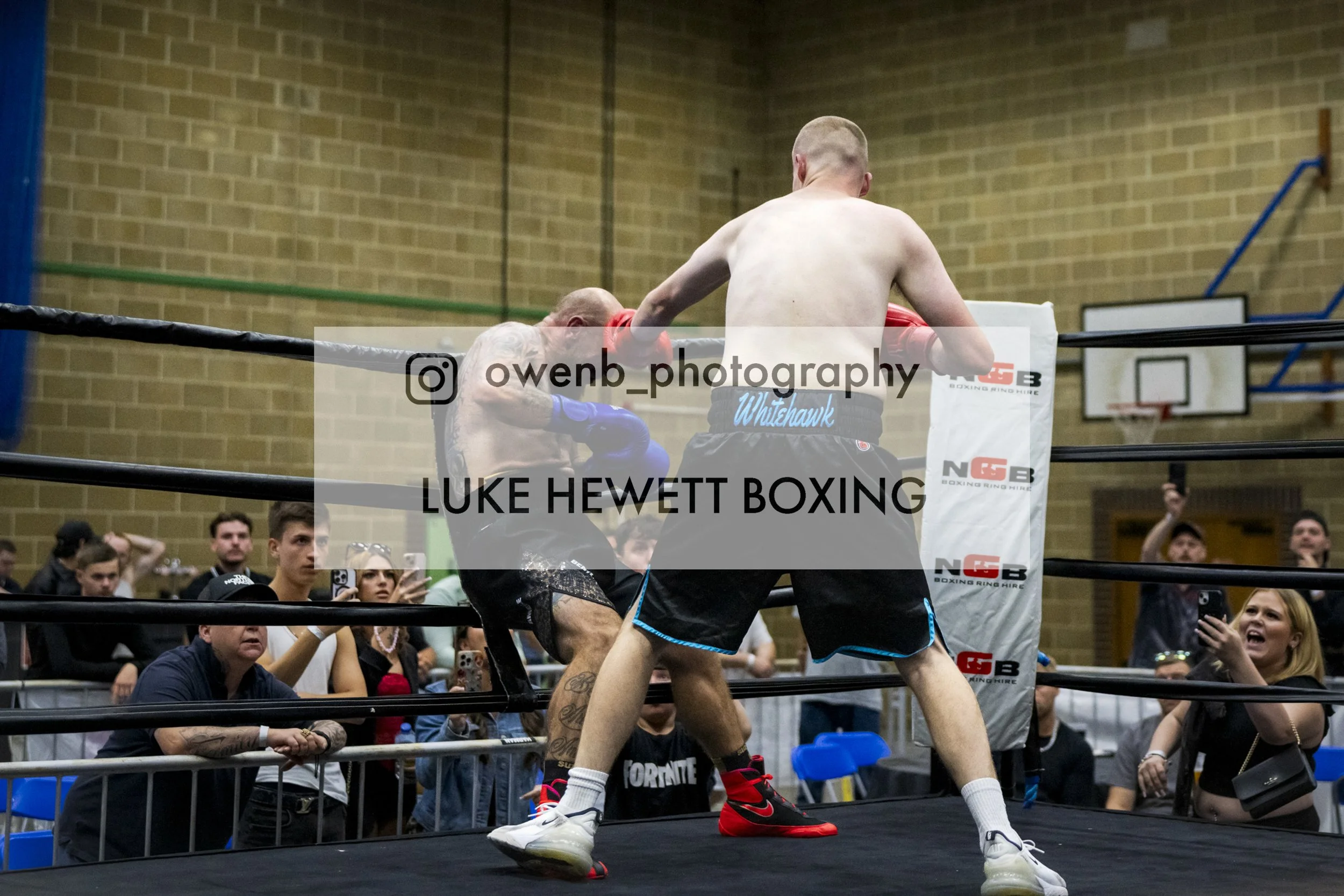Two male boxers are engaged in a match inside a boxing ring, with an audience watching around them. One boxer in black shorts is leaning against the corner pad while the other in white shorts with blue trim is delivering a punch. The background shows