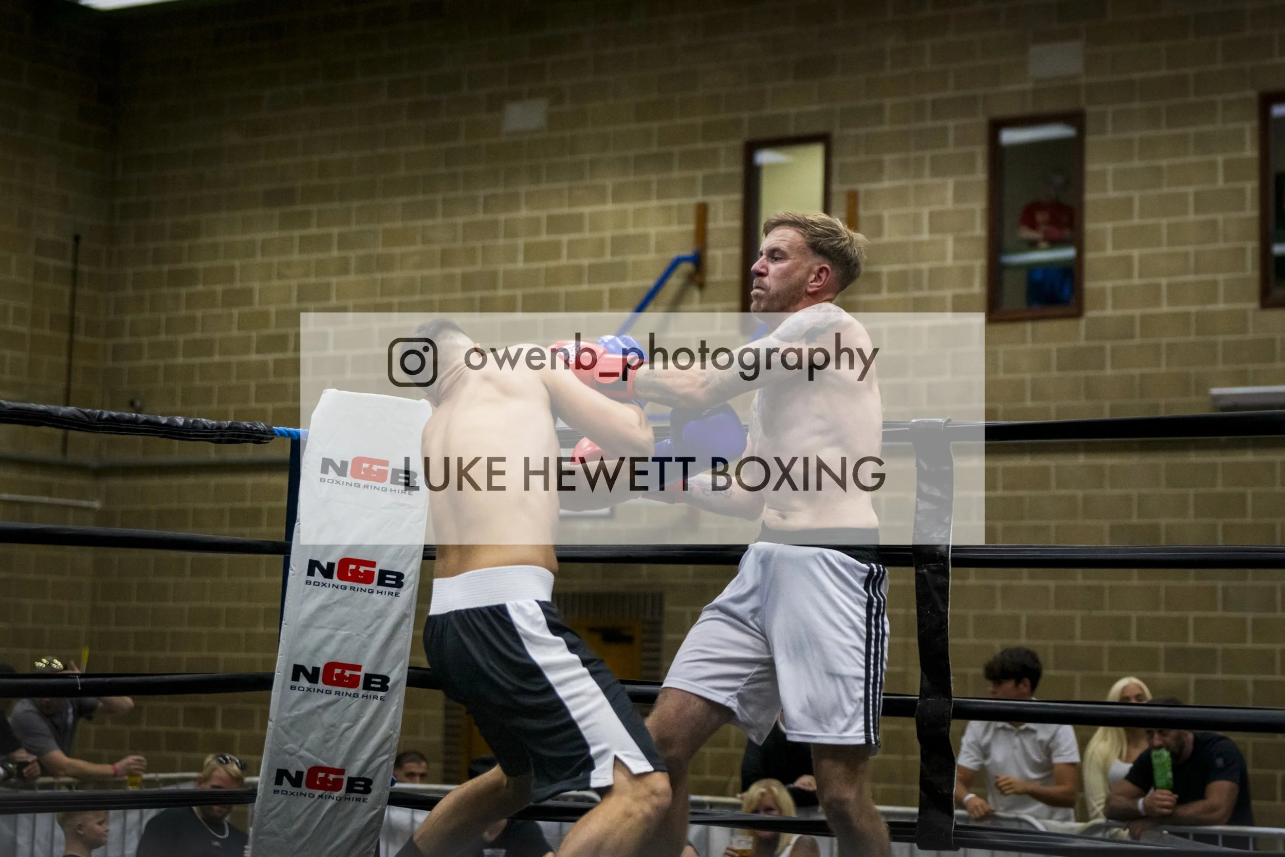 A boxing match with two fighters inside a ring, with one delivering a punch to the other, surrounded by spectators in a gymnasium.