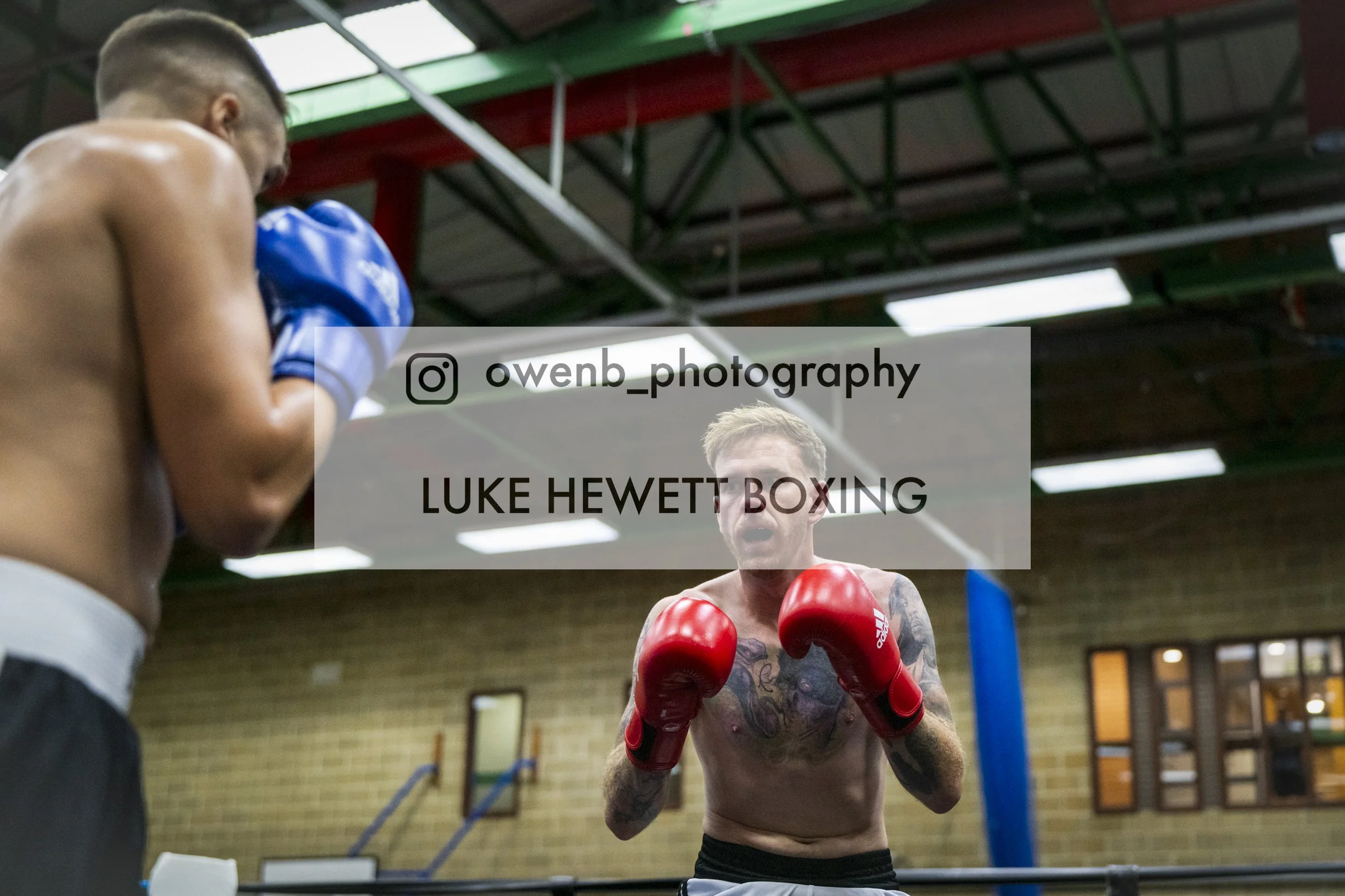 A shirtless male boxer with tattoos on his chest and arms and wearing red boxing gloves, facing a person in a blue training glove inside a boxing gym with a high ceiling and gym equipment.