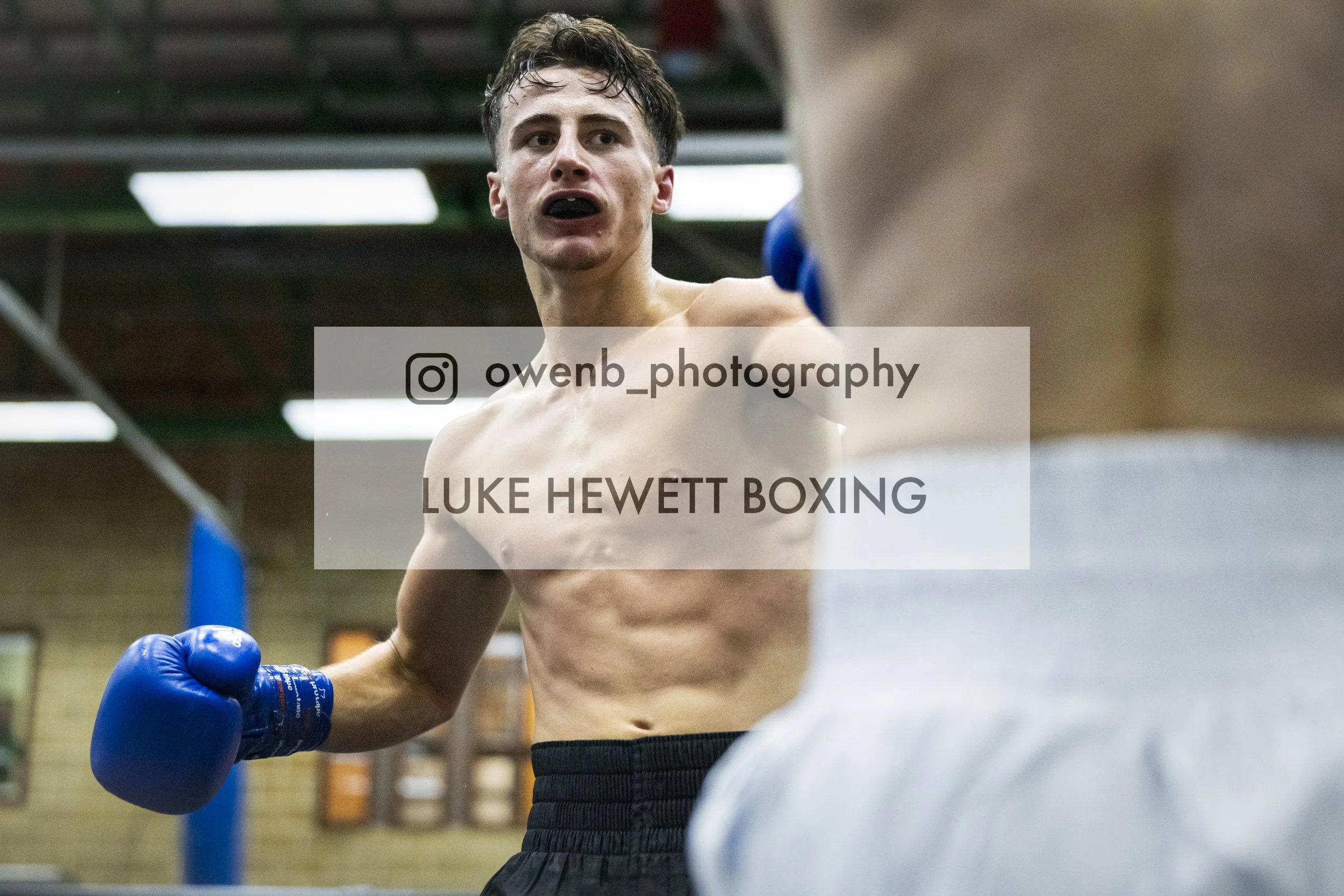 A shirtless male boxer wearing blue gloves in a gym, preparing for a boxing match with a trainer or opponent partially visible in the foreground.