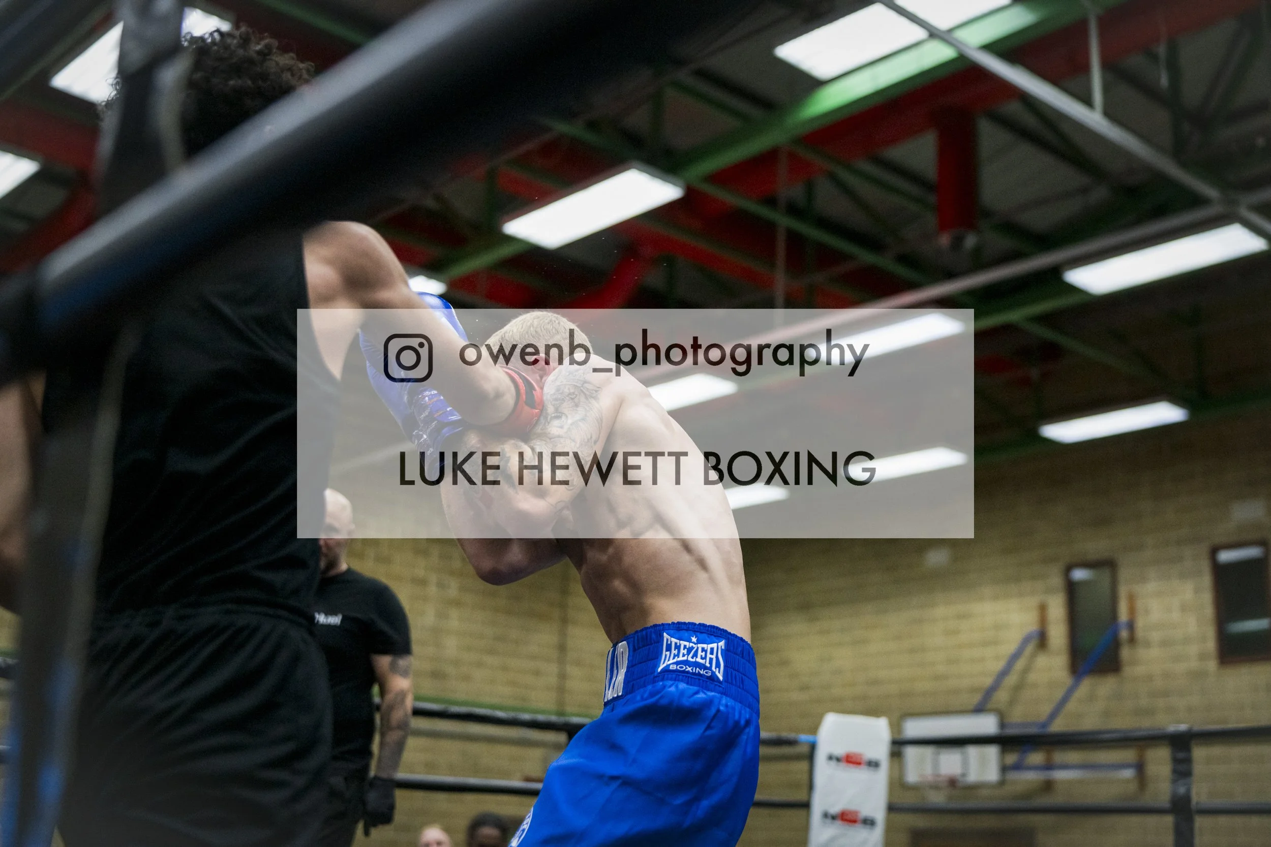 A boxer wearing blue shorts and gloves in a boxing ring, training or fighting, with training equipment and a gymnasium roof visible in the background.