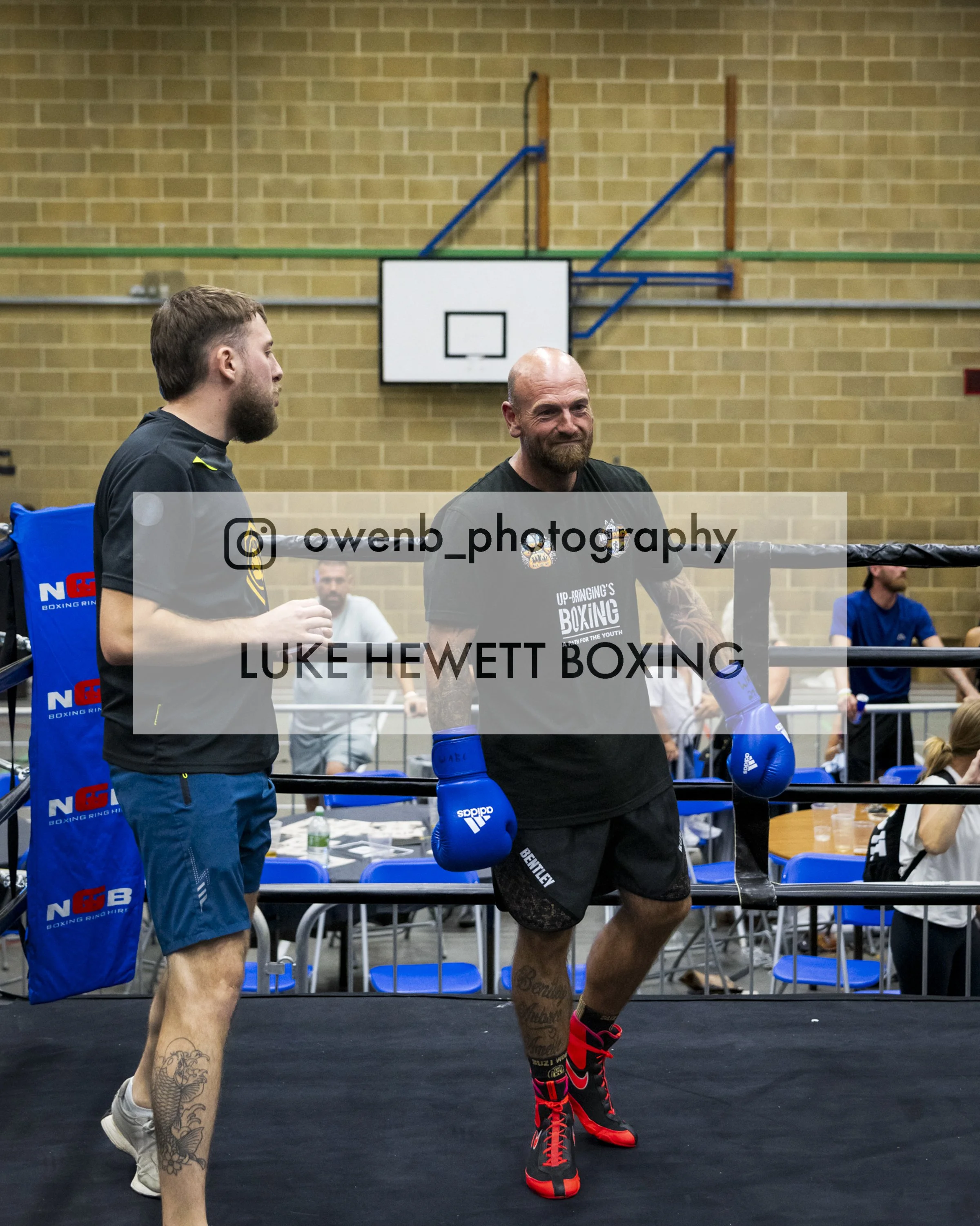 Two male boxers in boxing gear inside a gym or boxing ring. One is wearing black shorts and gloves, the other wearing navy shorts and gloves. They are preparing for a boxing match or training session, with a brick wall and gym equipment in the backgr