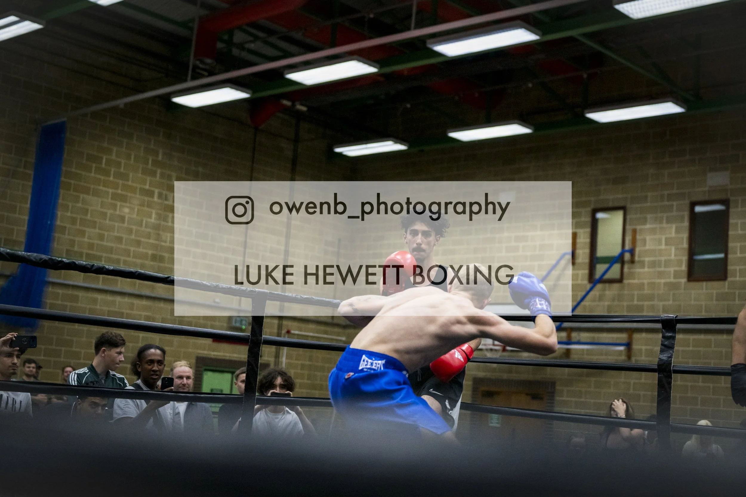 Boxer in blue shorts tackling an opponent over the ropes in a boxing ring during a match, with spectators watching in the background.
