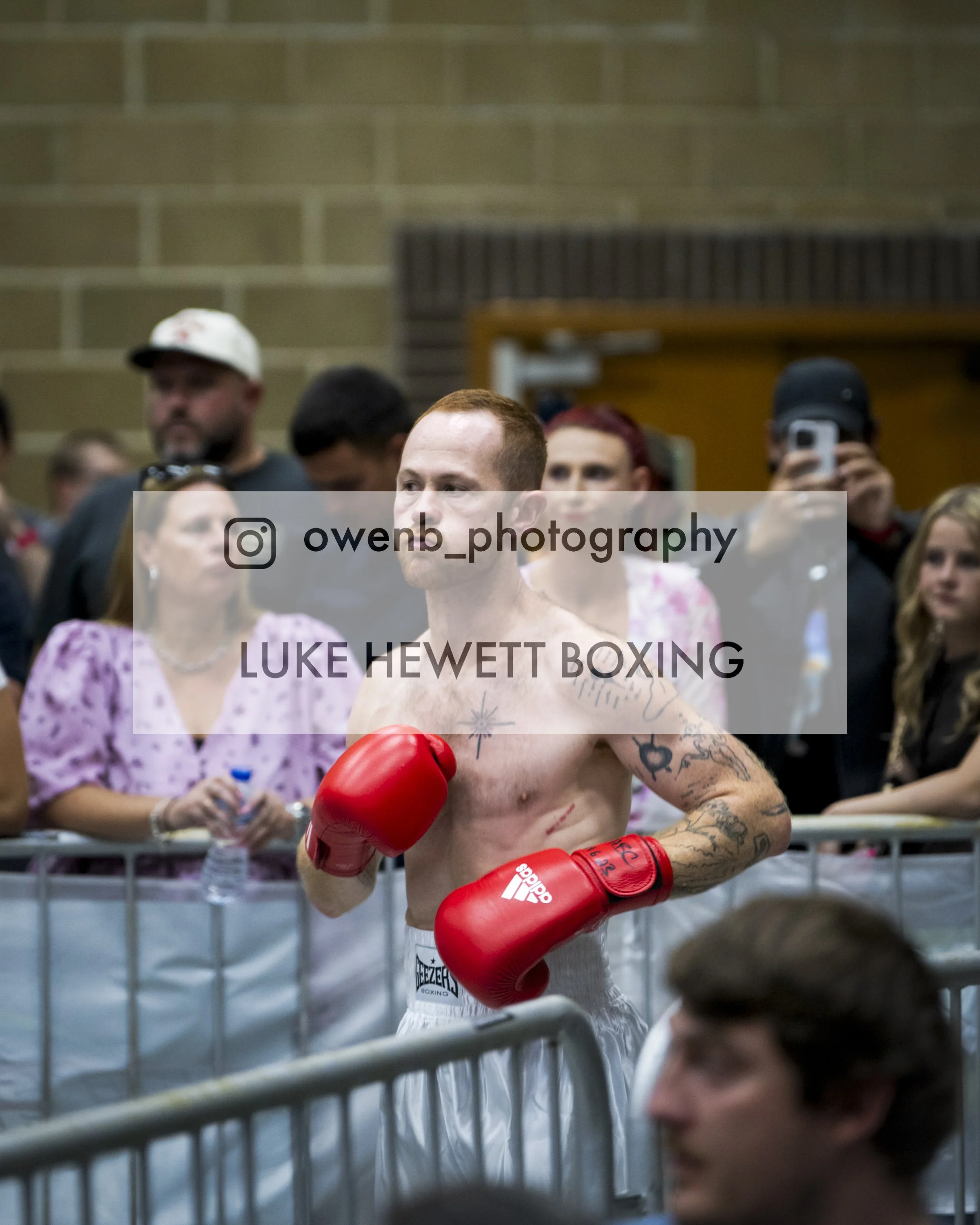 A shirtless male boxer with tattoos on his arm and chest, wearing red boxing gloves, standing inside a boxing ring with spectators behind the ropes. The crowd includes a woman in a pink floral dress, people taking photos, and others watching.