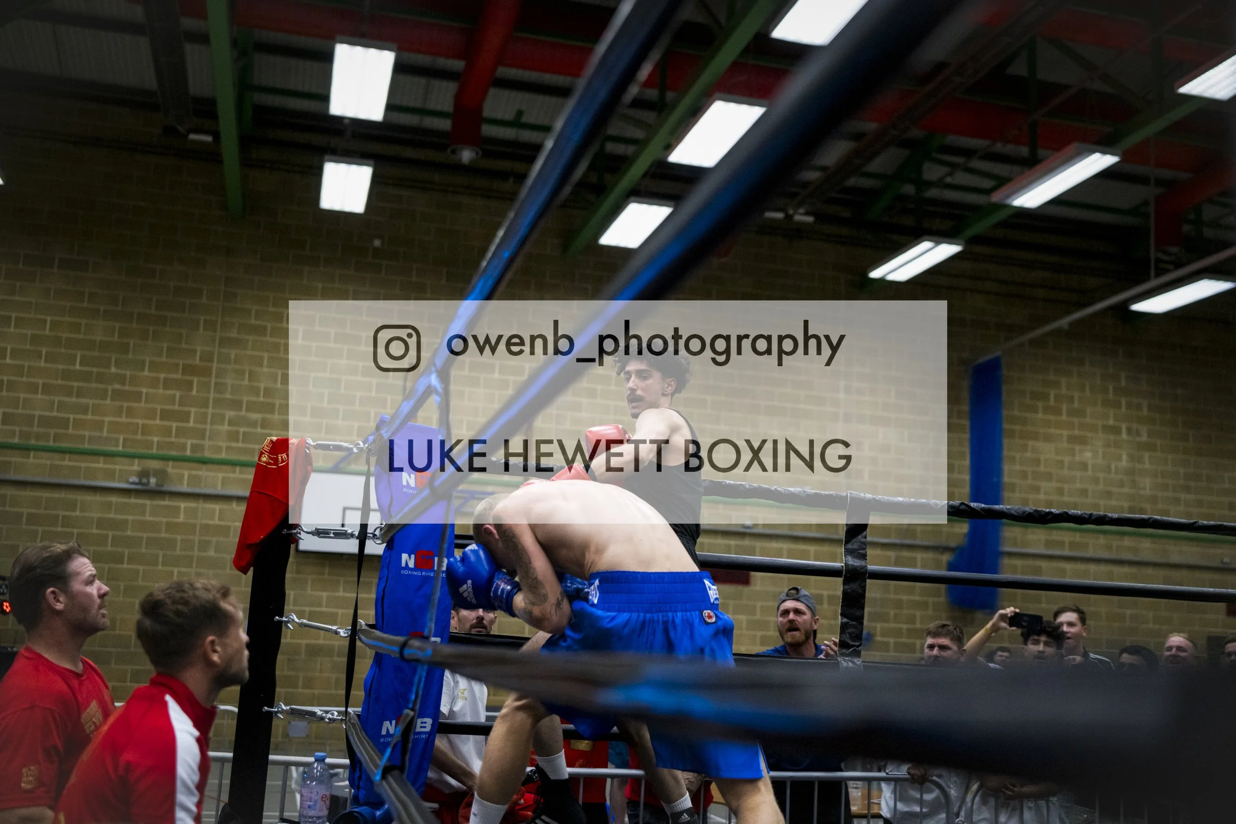 Boxers in a match inside a boxing ring surrounded by spectators, with one boxer leaning over the ropes and a referee looking on.