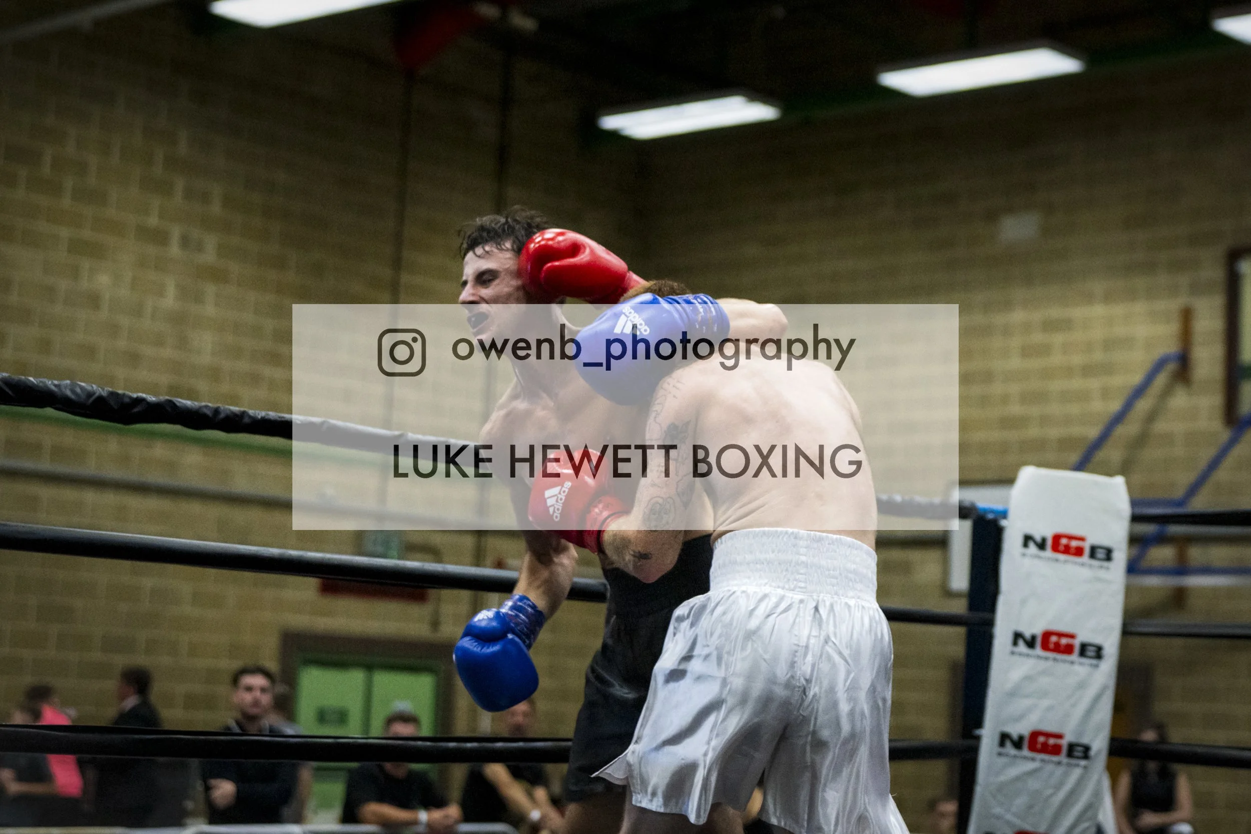 Boxer in a blue shirt delivers a punch to another boxer in white shorts inside a boxing ring. The scene is in a gym with a brick wall and onlookers in the background.