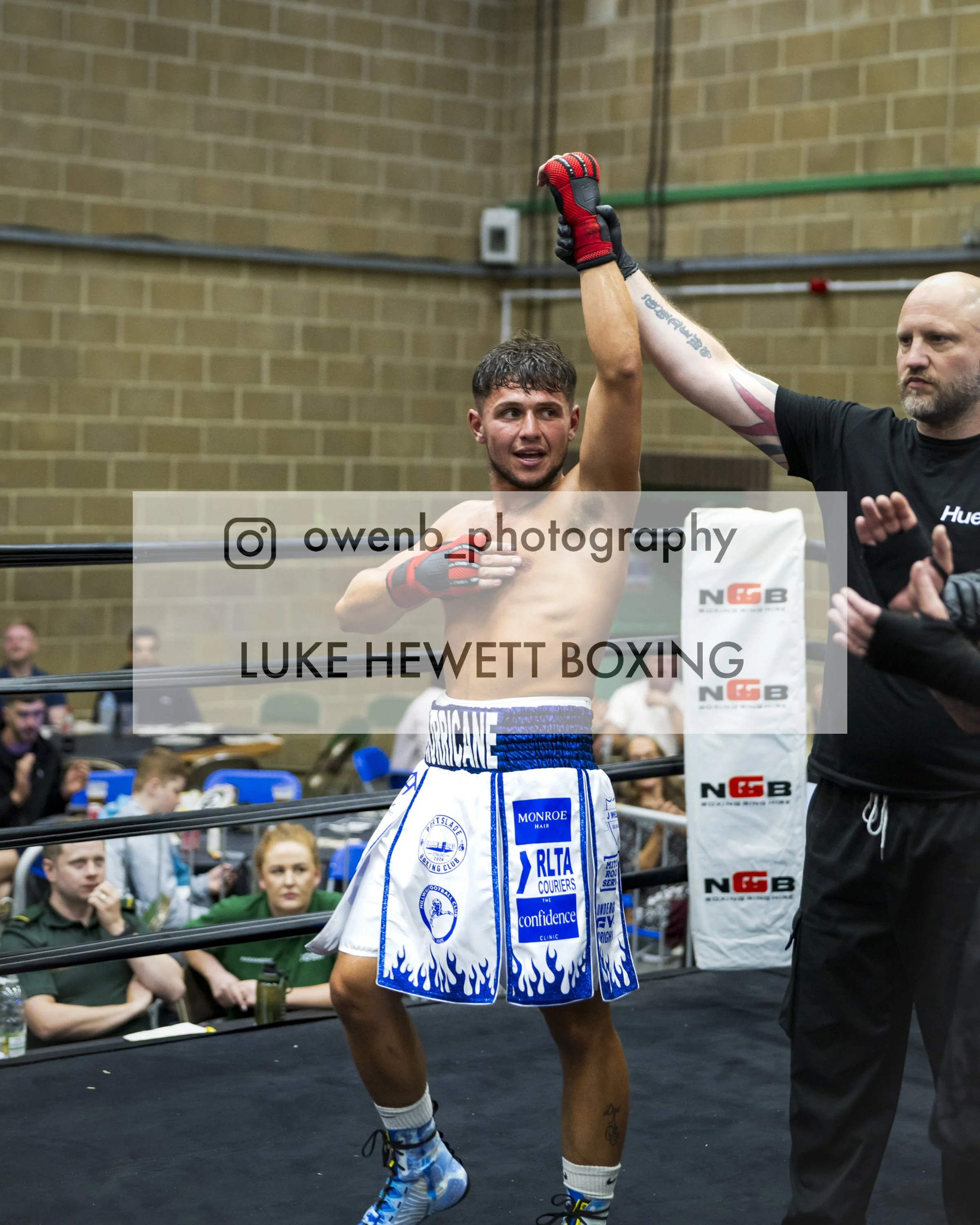 Boxer Luke Hewett raising his arm in victory inside a boxing ring, with an audience and a referee present, in a gymnasium with brick walls.