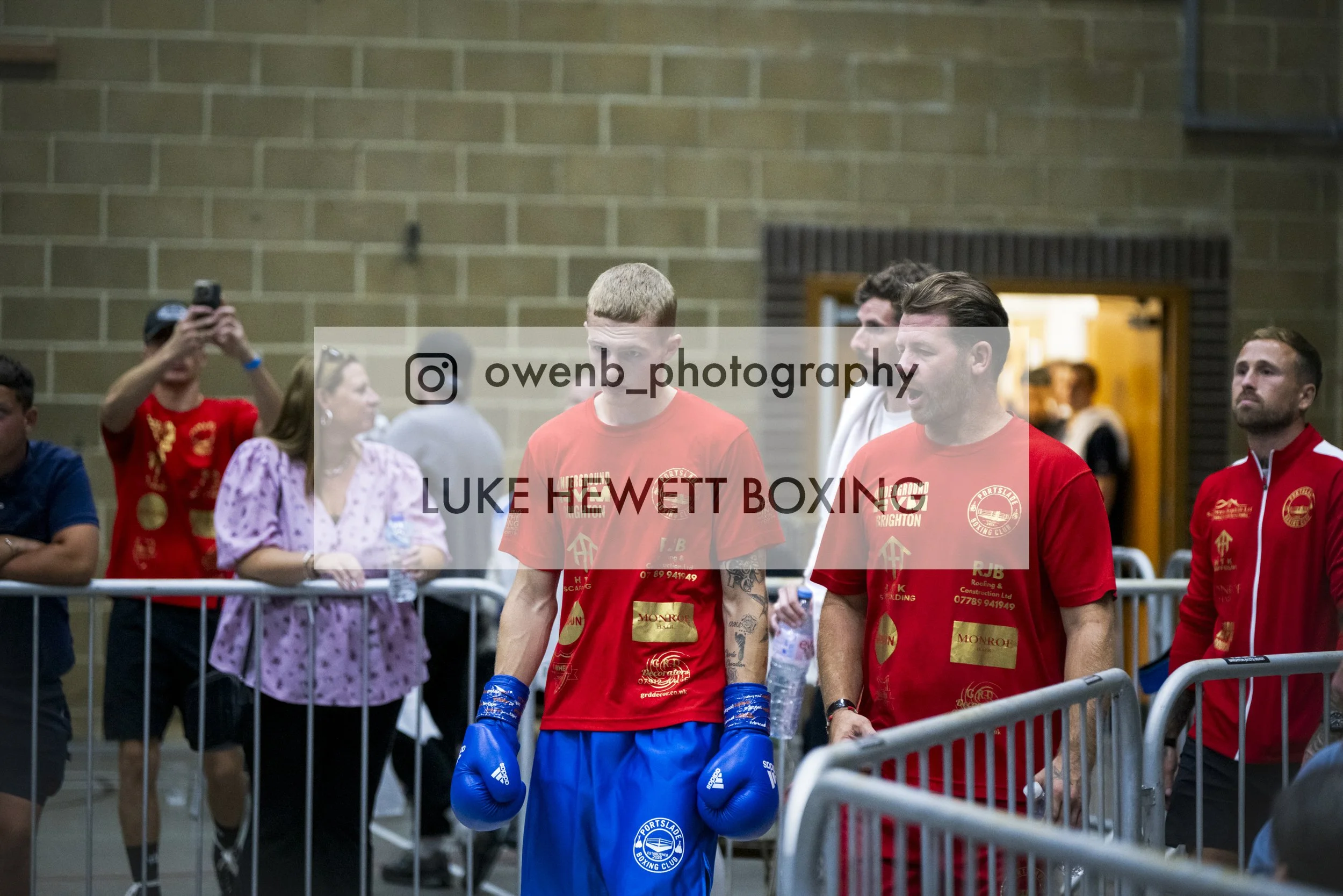 Boxer in blue gloves walking past spectators and officials inside a gymnasium.