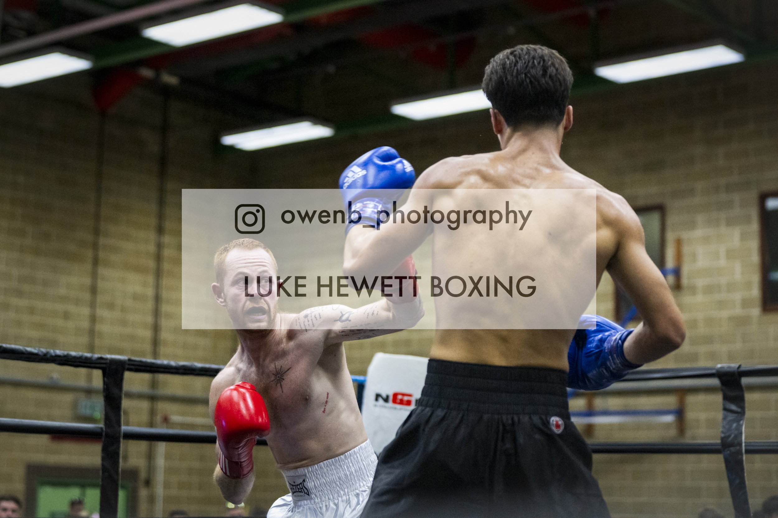 Two male boxers are engaged in a match inside a boxing ring. One is shirtless with boxing gloves, while the other is wearing a shirt and boxing gloves. The boxer without a shirt has a tattoo on his chest and appears to be throwing a punch, while the 