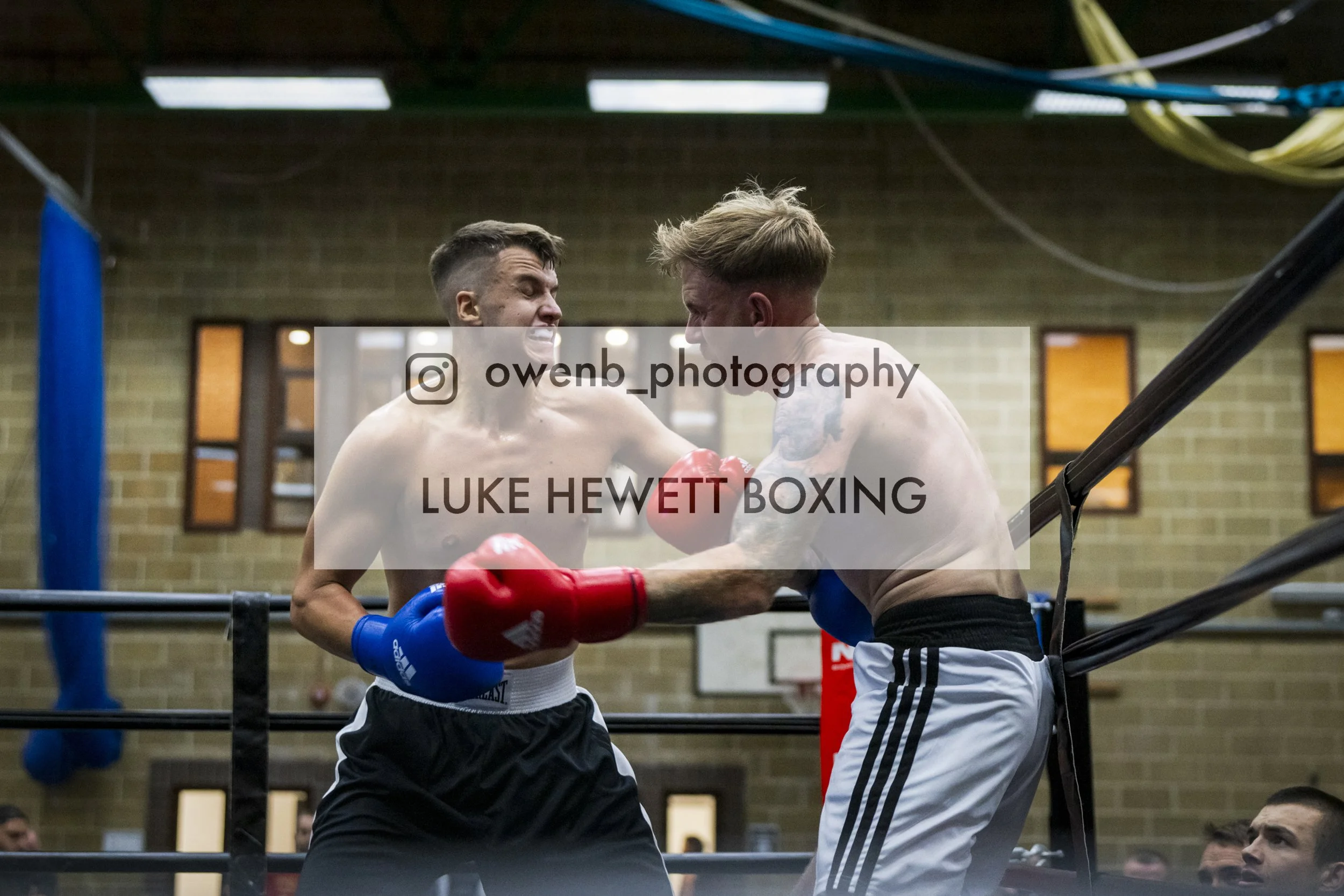 Two shirtless male boxers in gloves fighting inside a boxing ring with a brick wall background. One boxer throws a punch while the other reacts defensively, with audience members visible in the background.