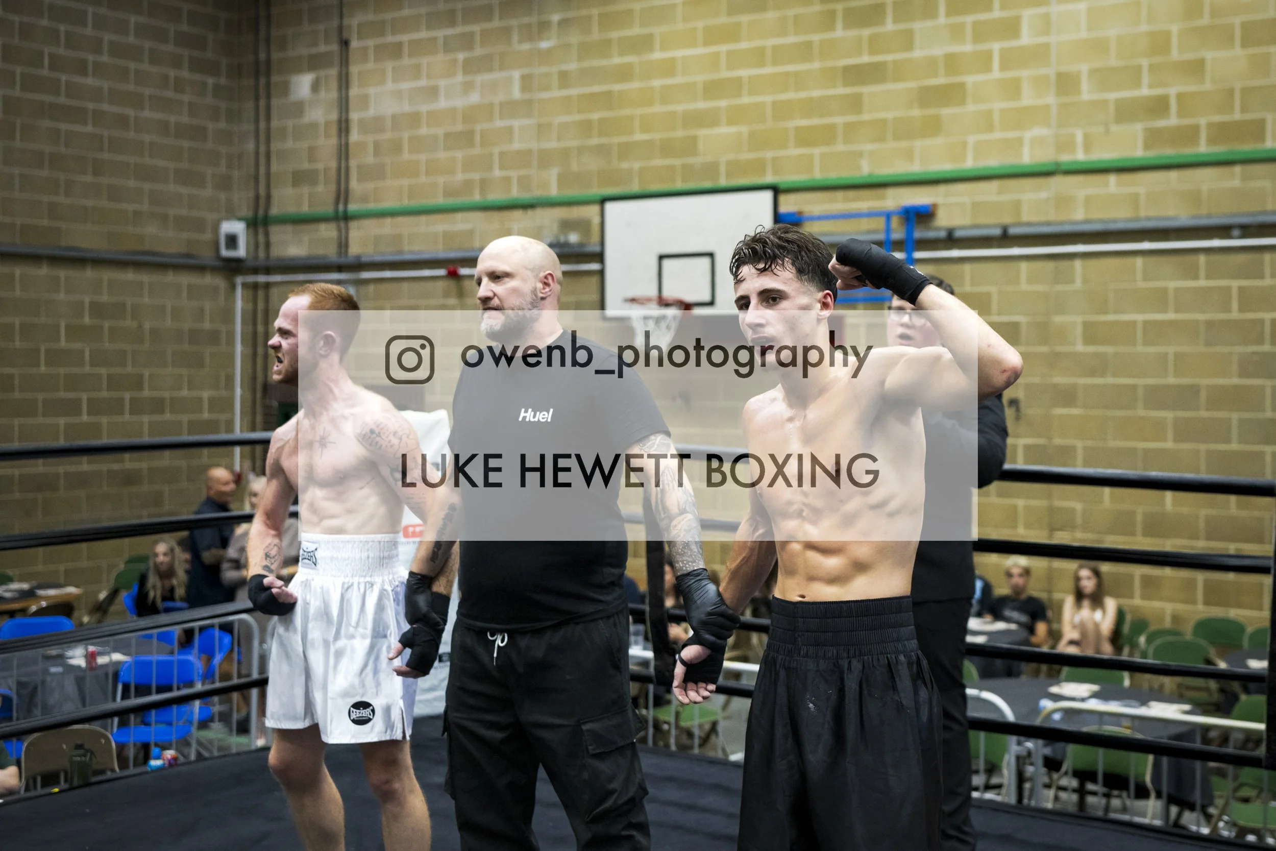 Two shirtless male boxers, one with red hair and tattoos and the other with dark curly hair, are in a boxing ring with their trainer behind them. The trainer and boxer have serious expressions, and the boxer with dark hair is adjusting his glove. The