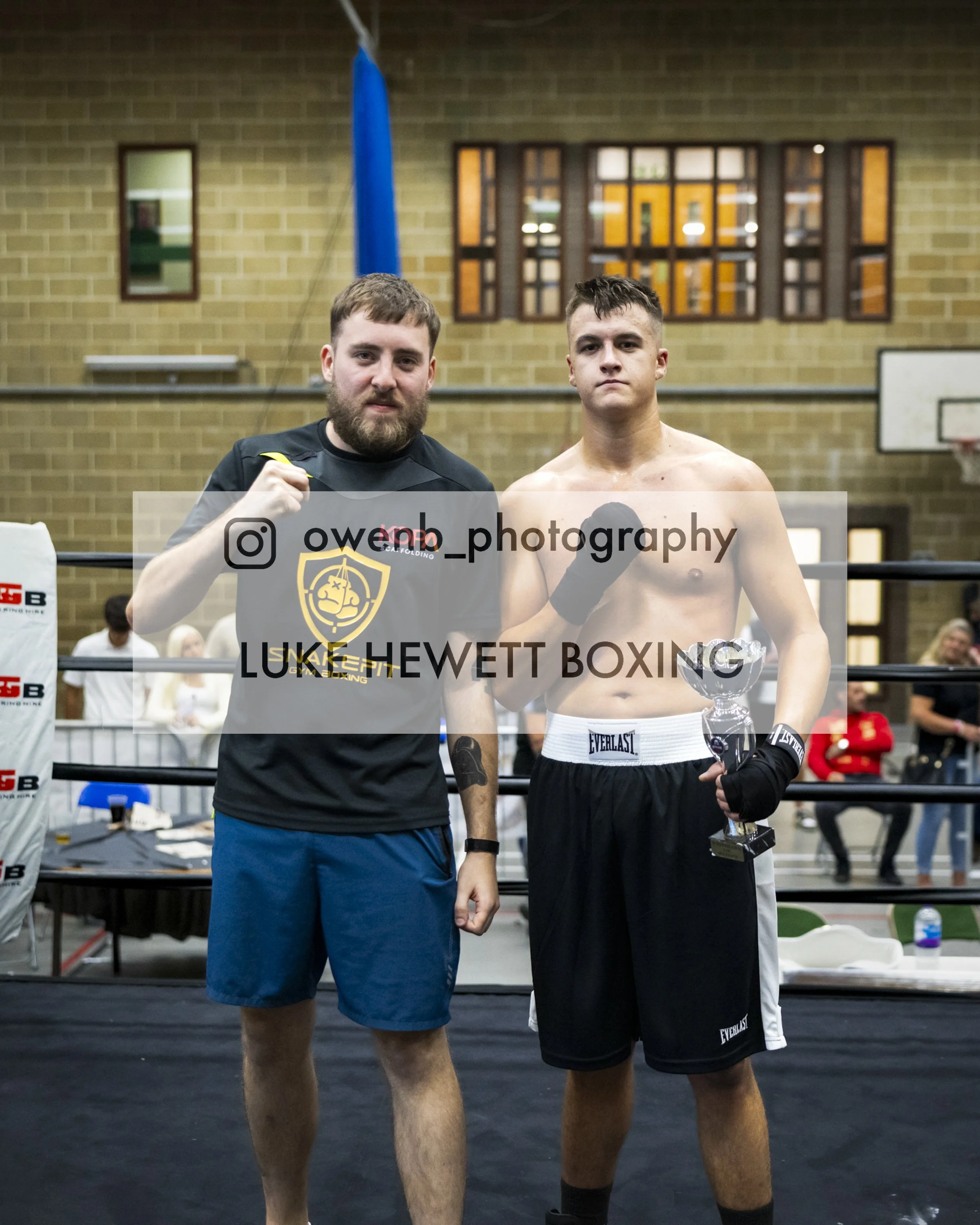 Two male boxers stand in a boxing ring, one is shirtless holding a trophy, the other is wearing a shirt with a logo and flexing his arm, both look serious, with a brick wall and a window in the background.