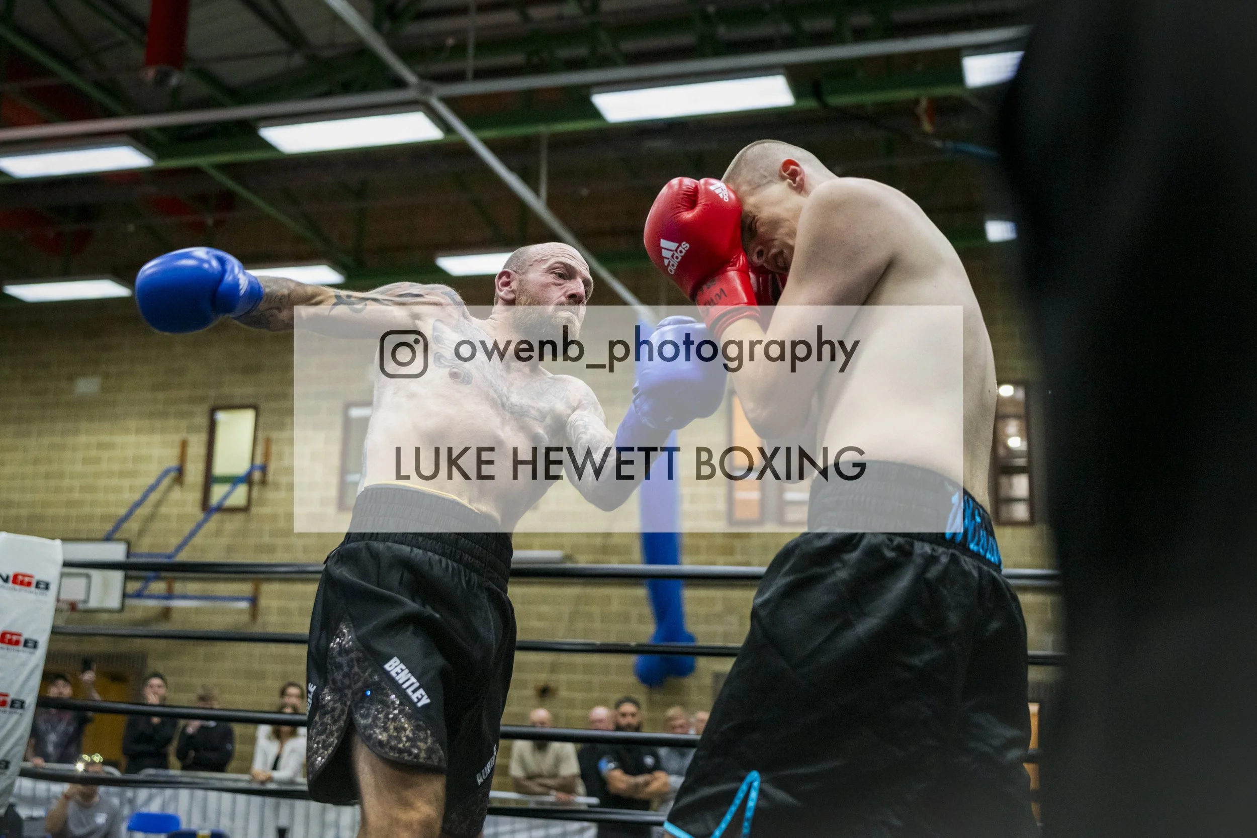 Two men with muscular builds and tattoos are boxing in a gym, one wearing blue gloves and the other wearing red gloves. They are engaged in a close exchange of punches, with the man in blue extending his punch toward the man with red gloves, who is l
