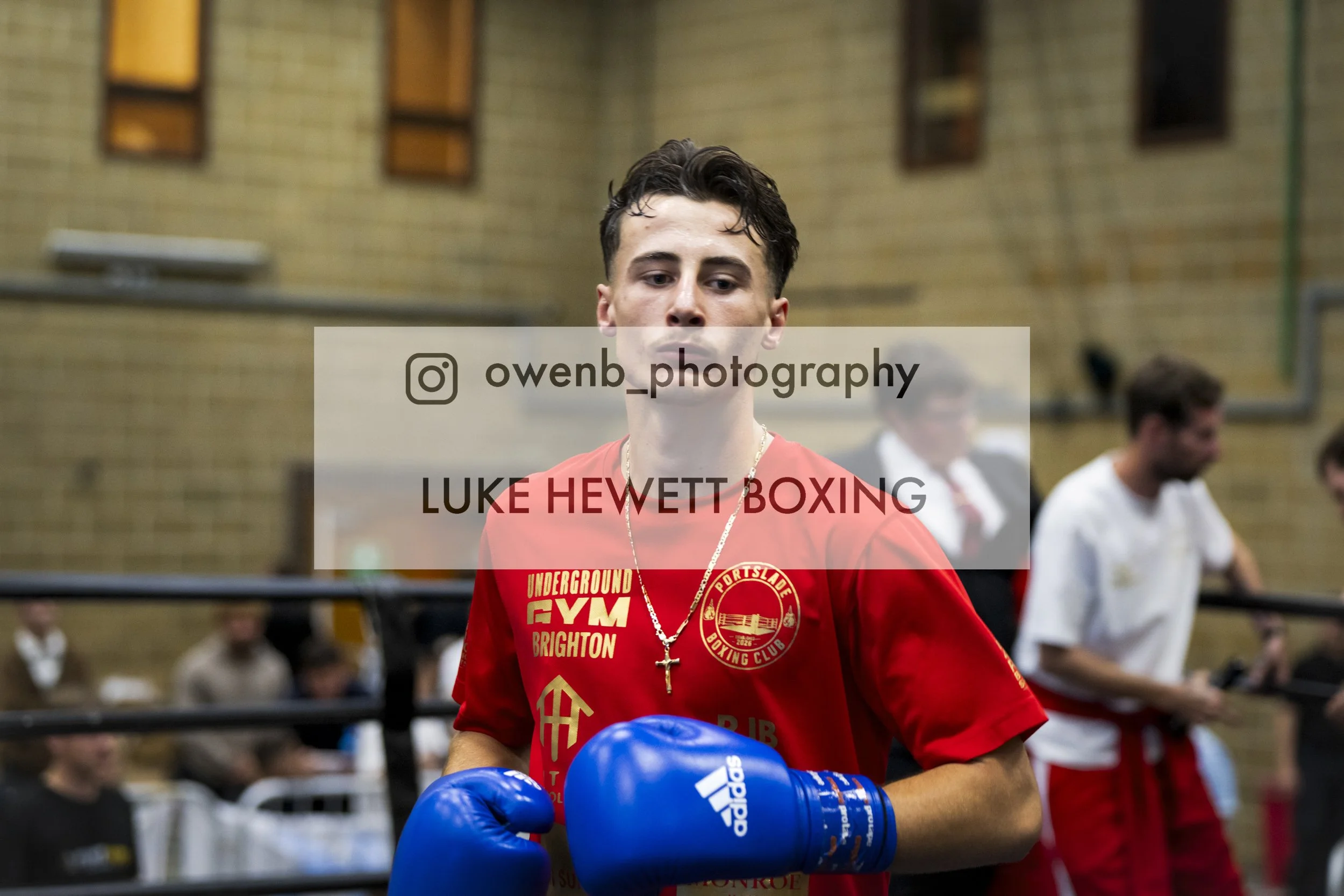 Young male boxer in red fight gear, wearing blue boxing gloves, standing in a boxing gym with a brick wall background, and people sitting in the background.