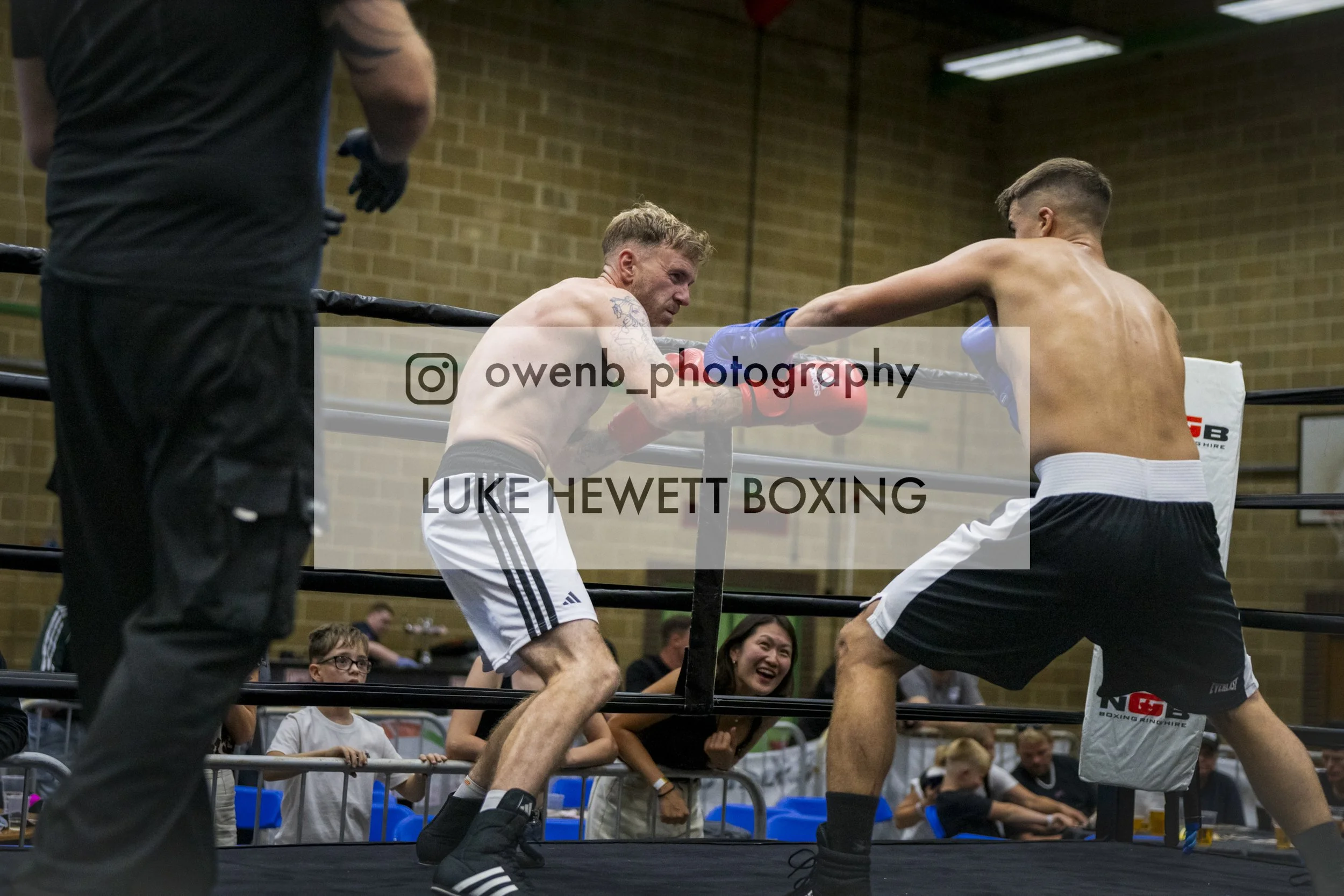 Two shirtless male boxers engaged in a match inside a boxing ring. One is throwing a punch while the other is leaning back. Several spectators are watching, some smiling. The scene takes place in a gymnasium with brick walls.
