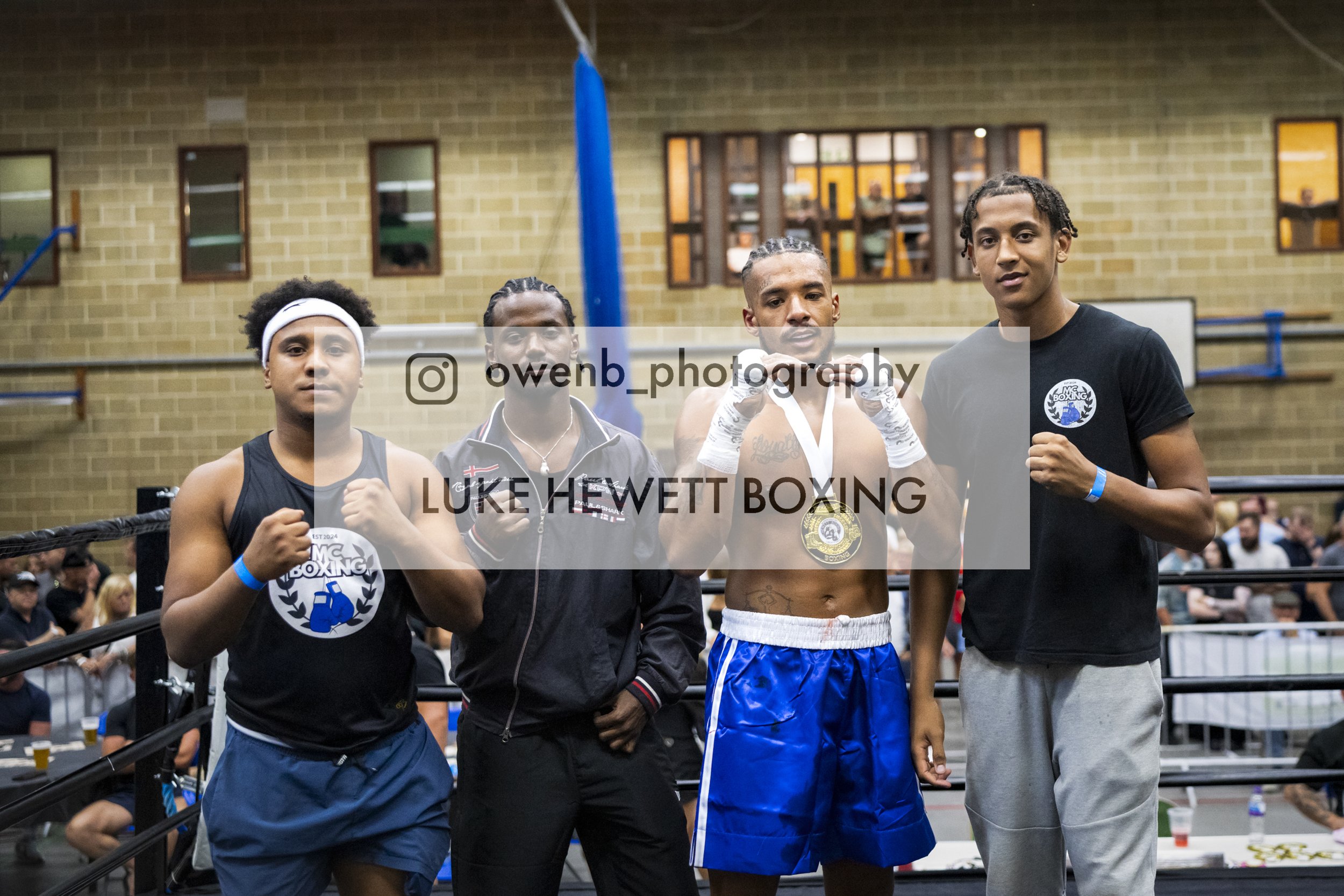Four young men pose in a boxing gym, standing side by side with some raising fists or fists in front of them. They are wearing athletic attire, and two are wearing medals around their necks. Behind them, there is a boxing ring and spectators. The gym