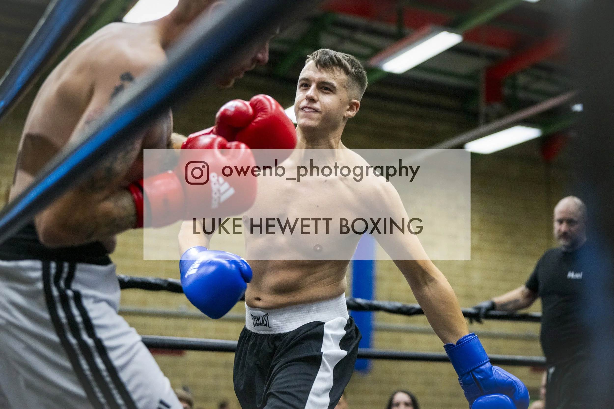 A shirtless male boxer with short dark hair and a focused expression, wearing blue gloves and black and white shorts, is being coached or guided by another person in a gym. The coach is holding red boxing gloves. The gym has a green and red ceiling w
