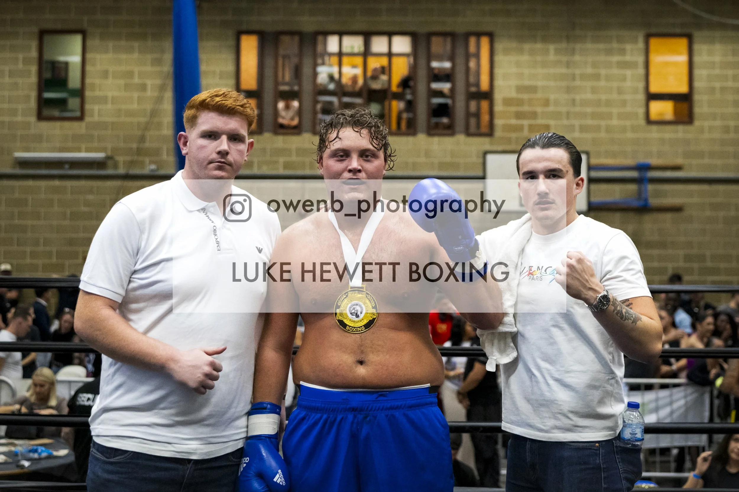Three men posing in a boxing ring after a match, with a boxer in the middle wearing blue shorts and gloves, holding a gold medal, flanked by two men in casual clothing, one wearing a white shirt and the other wearing a white shirt with tattoos.