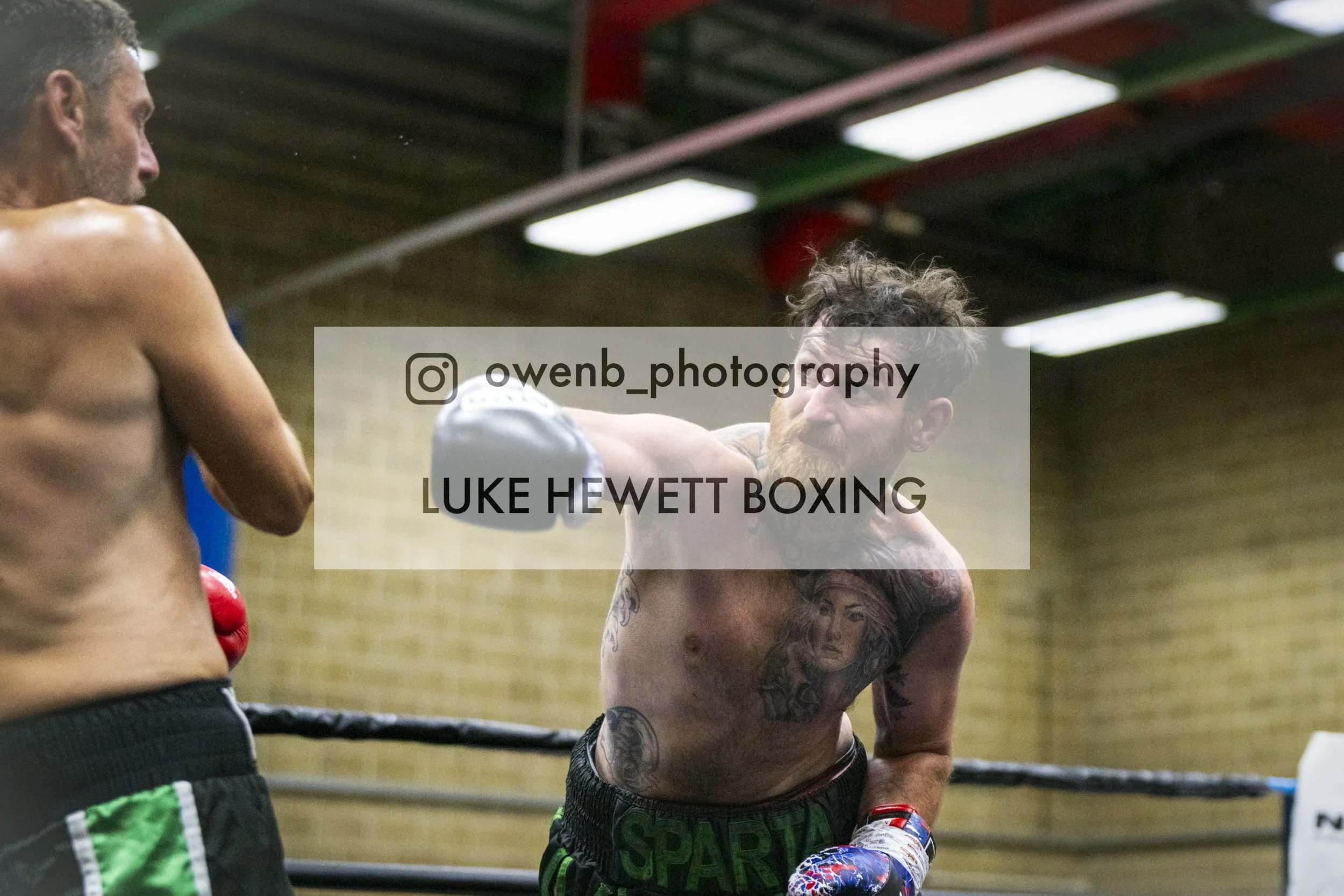 Two shirtless male boxers with gloves training in a boxing ring, one with a beard and tattoos, and the other with short hair, in a gym with a wooden ceiling.