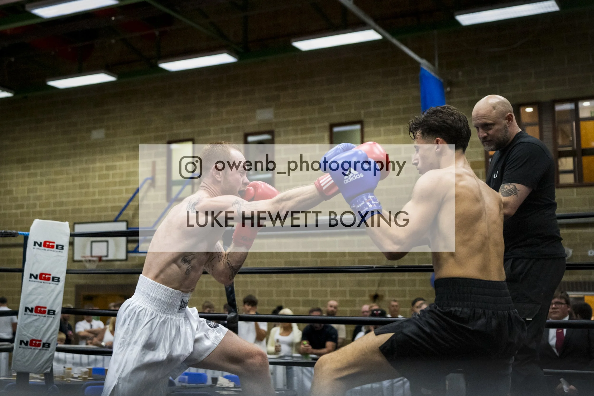 Two men engaged in a boxing match inside a gymnasium, with one man wearing red gloves and the other wearing blue gloves, and a referee or coach standing nearby.